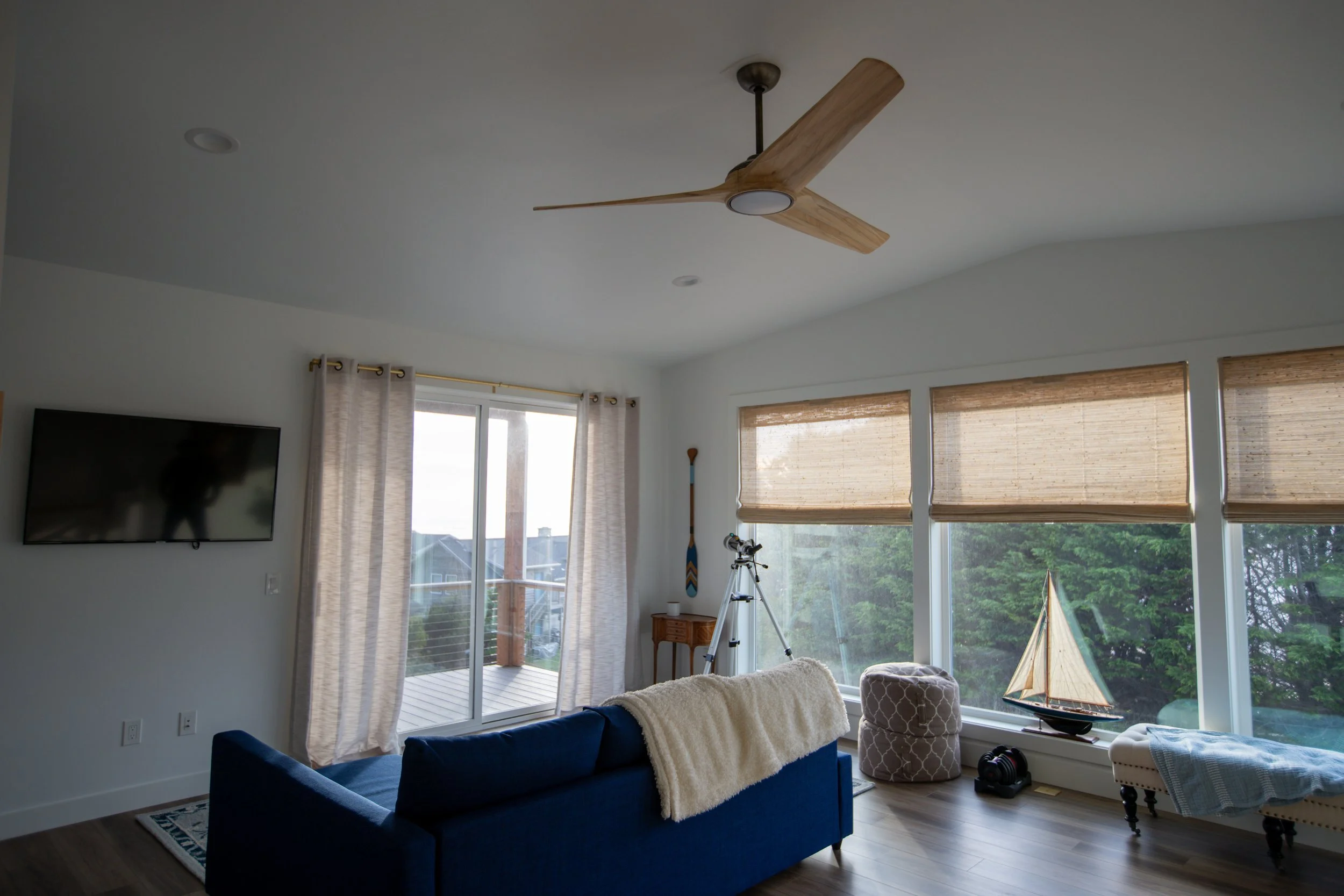 A living room with large windows, a sliding glass door, a blue sofa with a cream throw, a telescope, a model sailboat, a ceiling fan, and beige blinds and curtains.