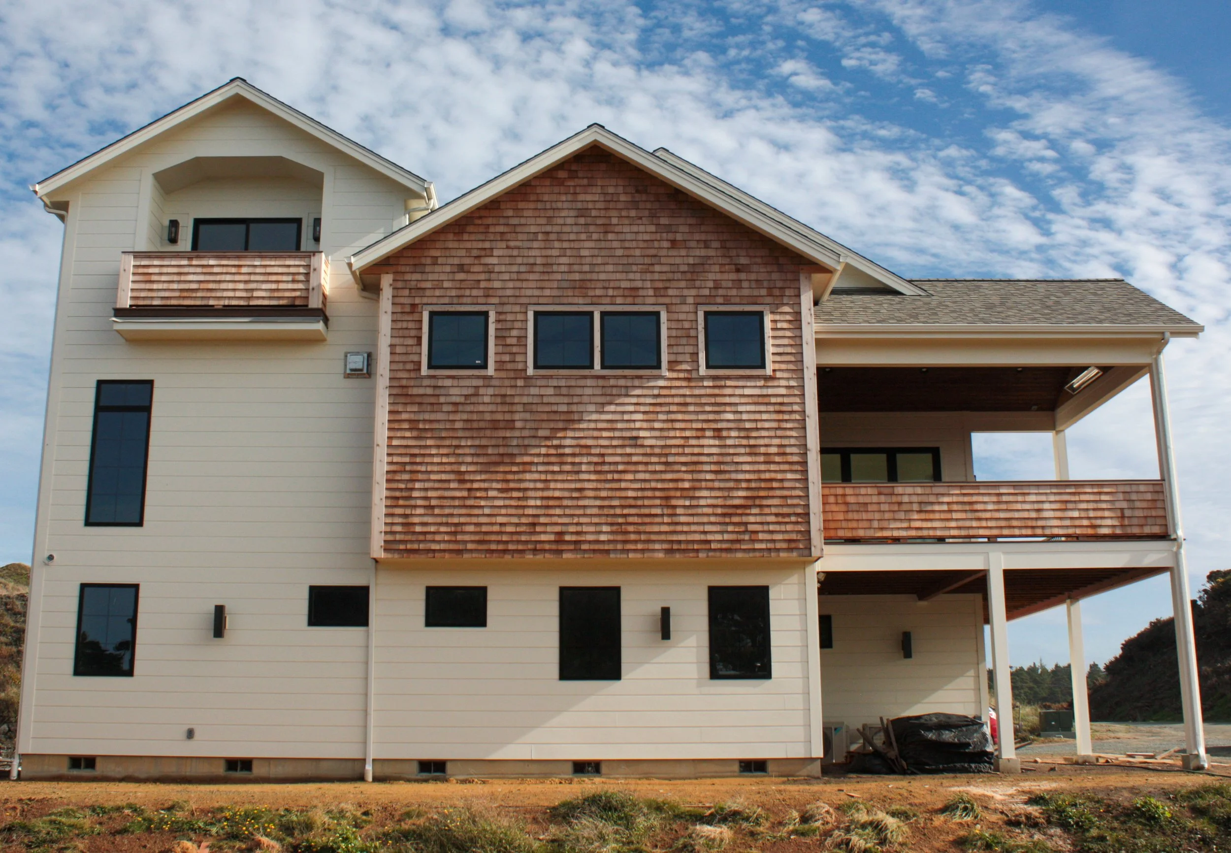 A modern, multi-story house under construction with white siding, brown shingled accents, and a balcony, set against a partly cloudy sky.