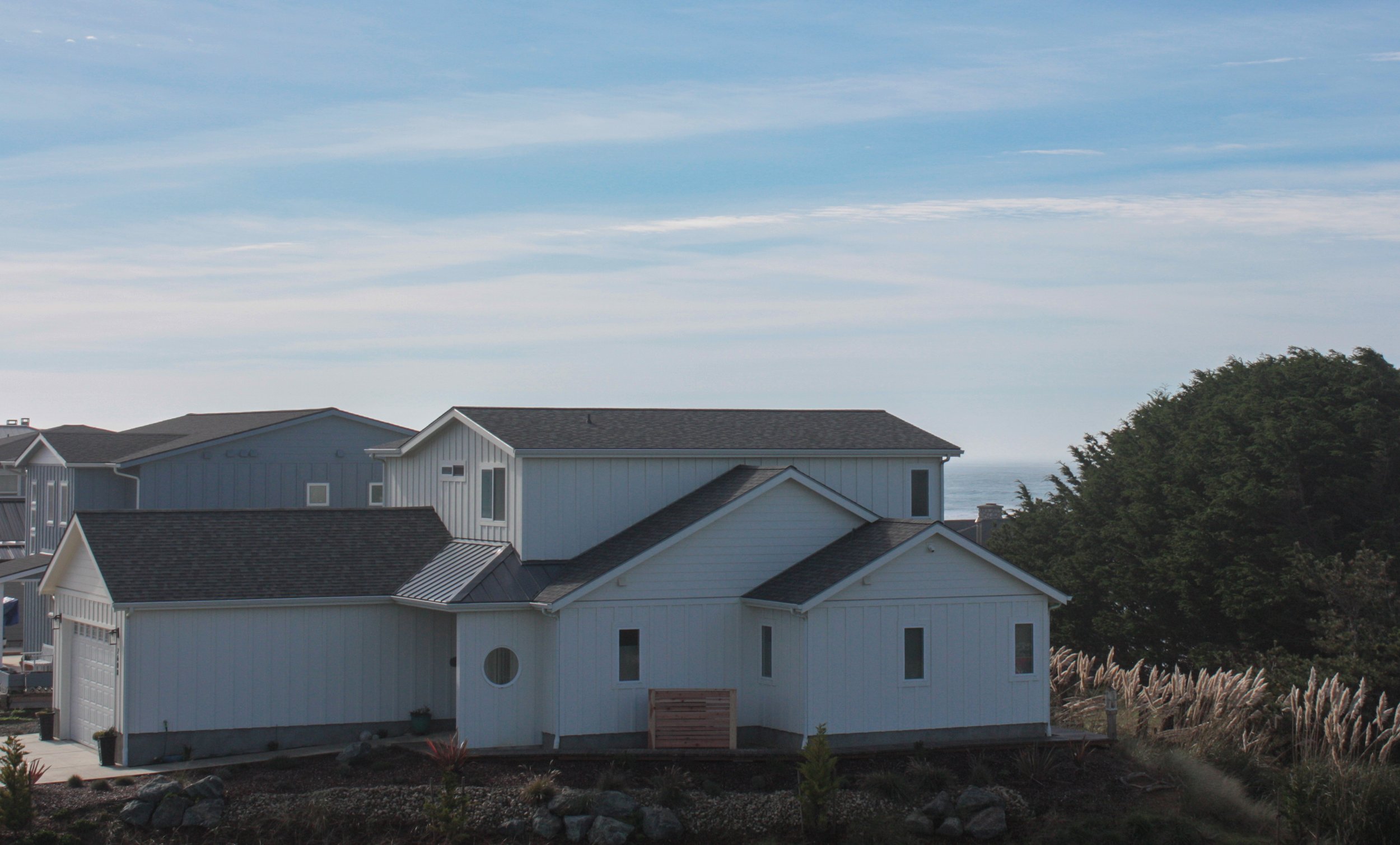 White Coastal-style house with multiple gable roofs, located near the ocean with greenery and a sky with wispy clouds in the background.
