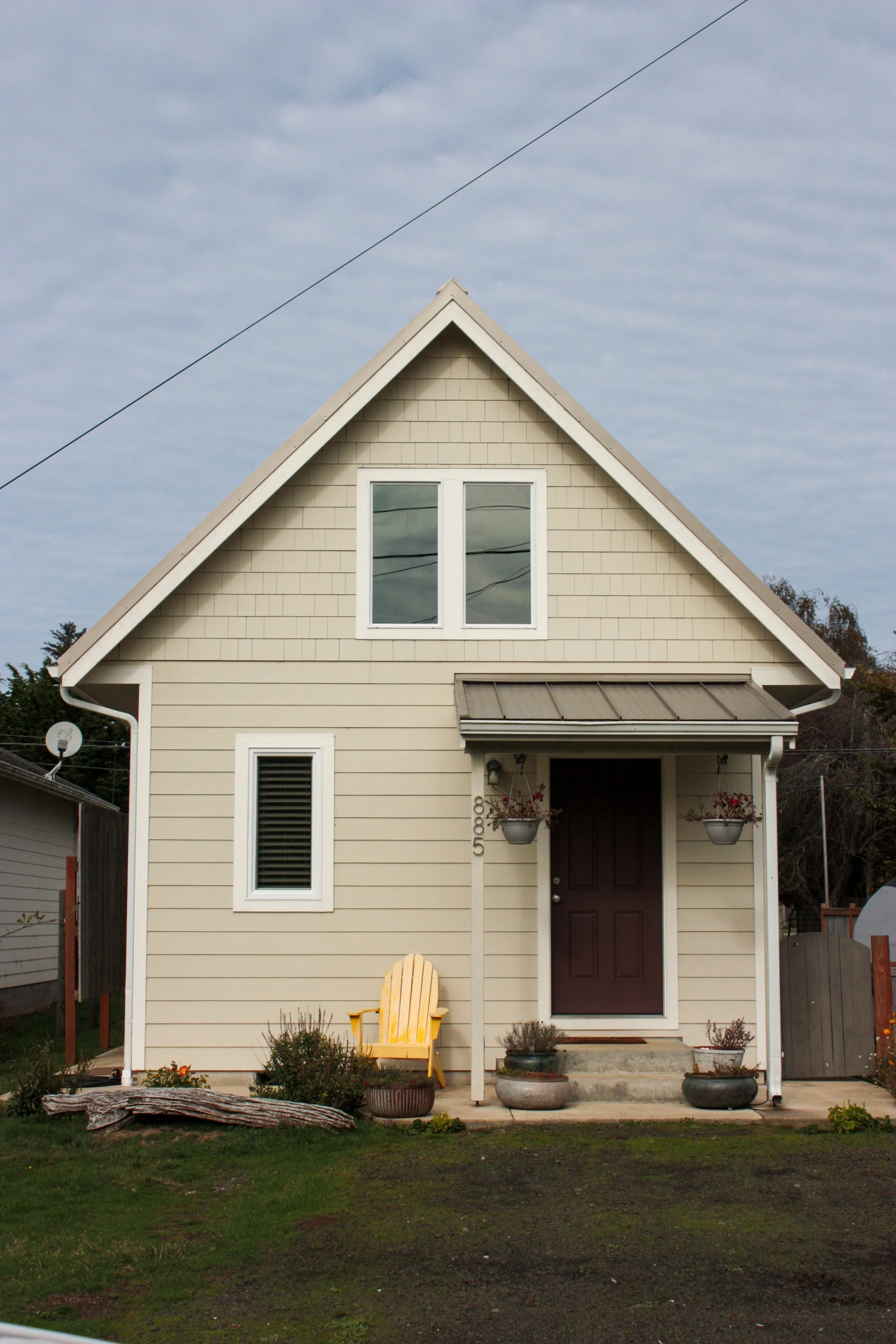 A beige house with a dark front door, two hanging flower baskets, a yellow outdoor chair, and several potted plants in front.