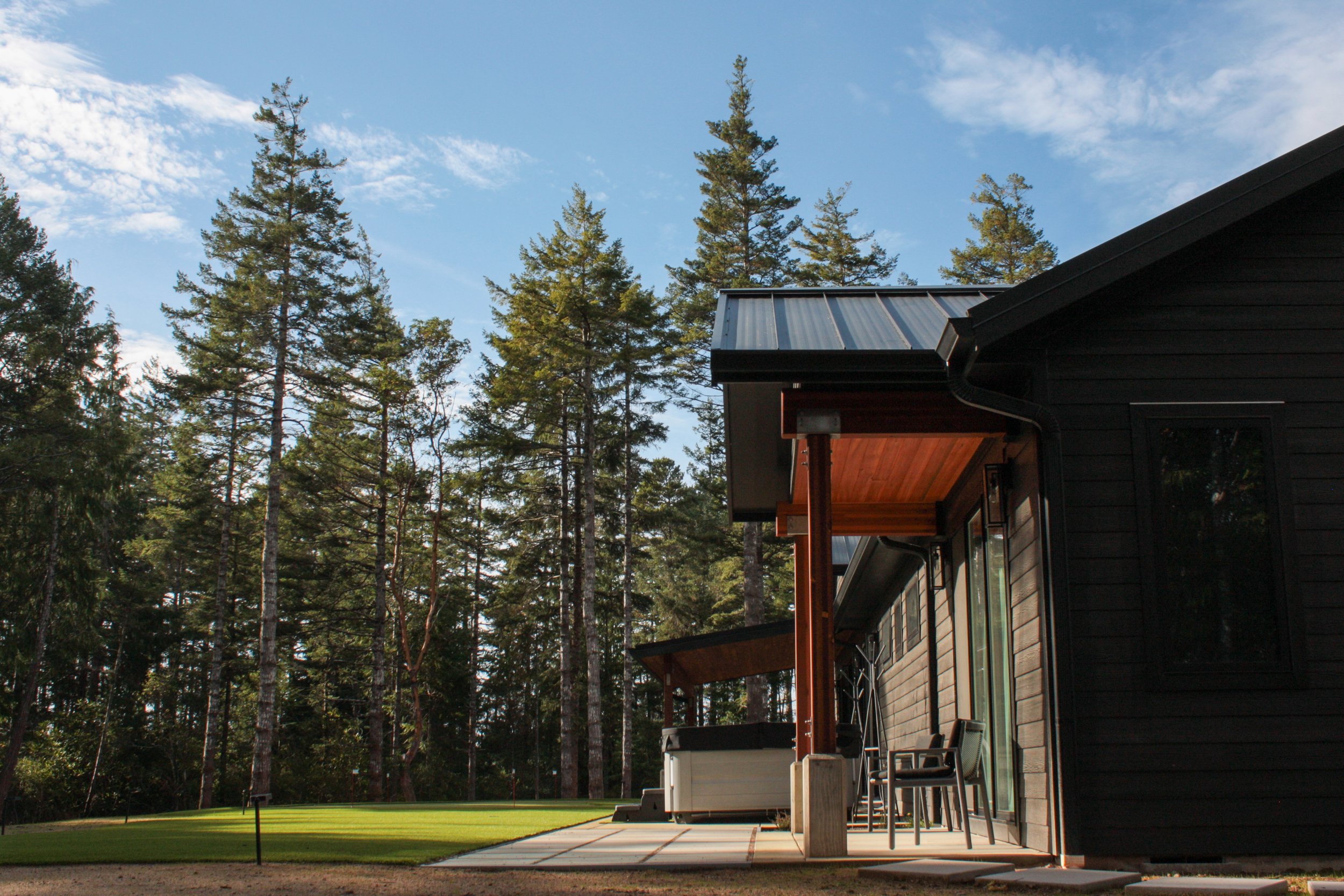 Back view of a black modern house with wooden accents, a patio with chairs, and a hot tub, surrounded by tall pine trees under a blue sky.