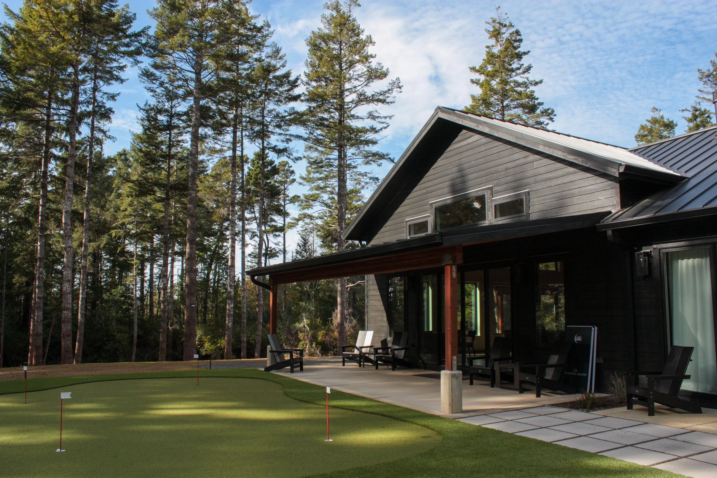 A modern house with a dark exterior and large windows, surrounded by a patio with chairs, adjacent to a golf course green with flags, and tall pine trees in the background under a partly cloudy sky.