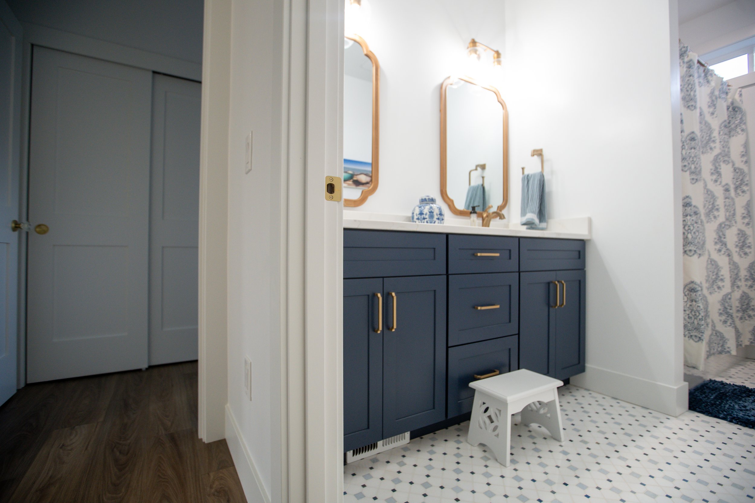 Bathroom with navy blue vanity, two mirrors, gold hardware, and a white step stool, with a patterned shower curtain to the right.