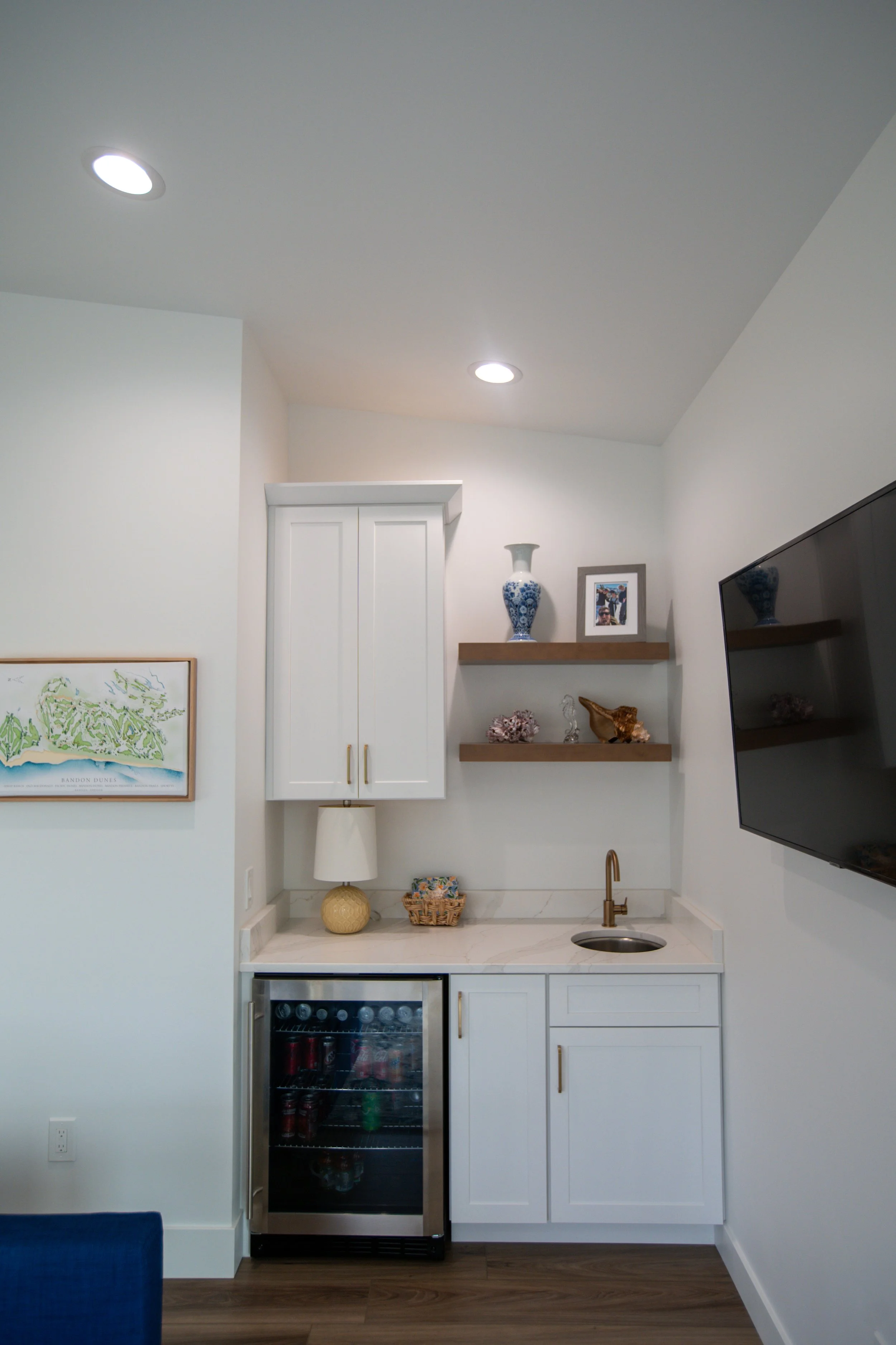 Mini fridge with glass door, white kitchen cabinets, a small sink with a gold faucet, wooden shelves with decorative items, a framed photo, a table lamp, and a wall art map of Bandon Dunes golf resort.