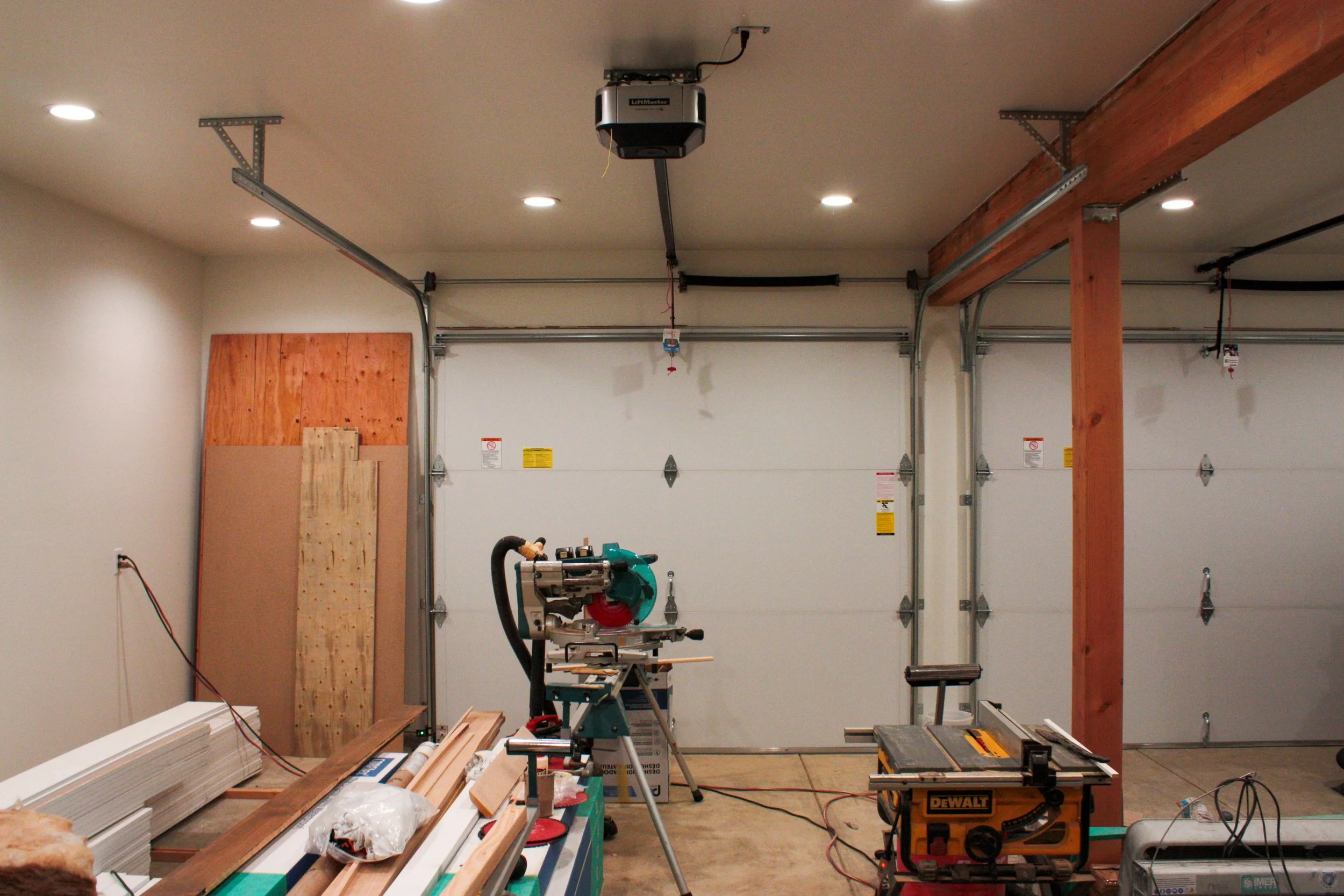 Garage with tools and construction materials, including a saw, wood planks, and a partially finished wooden beam. The garage door is closed. There are overhead lights on the ceiling.