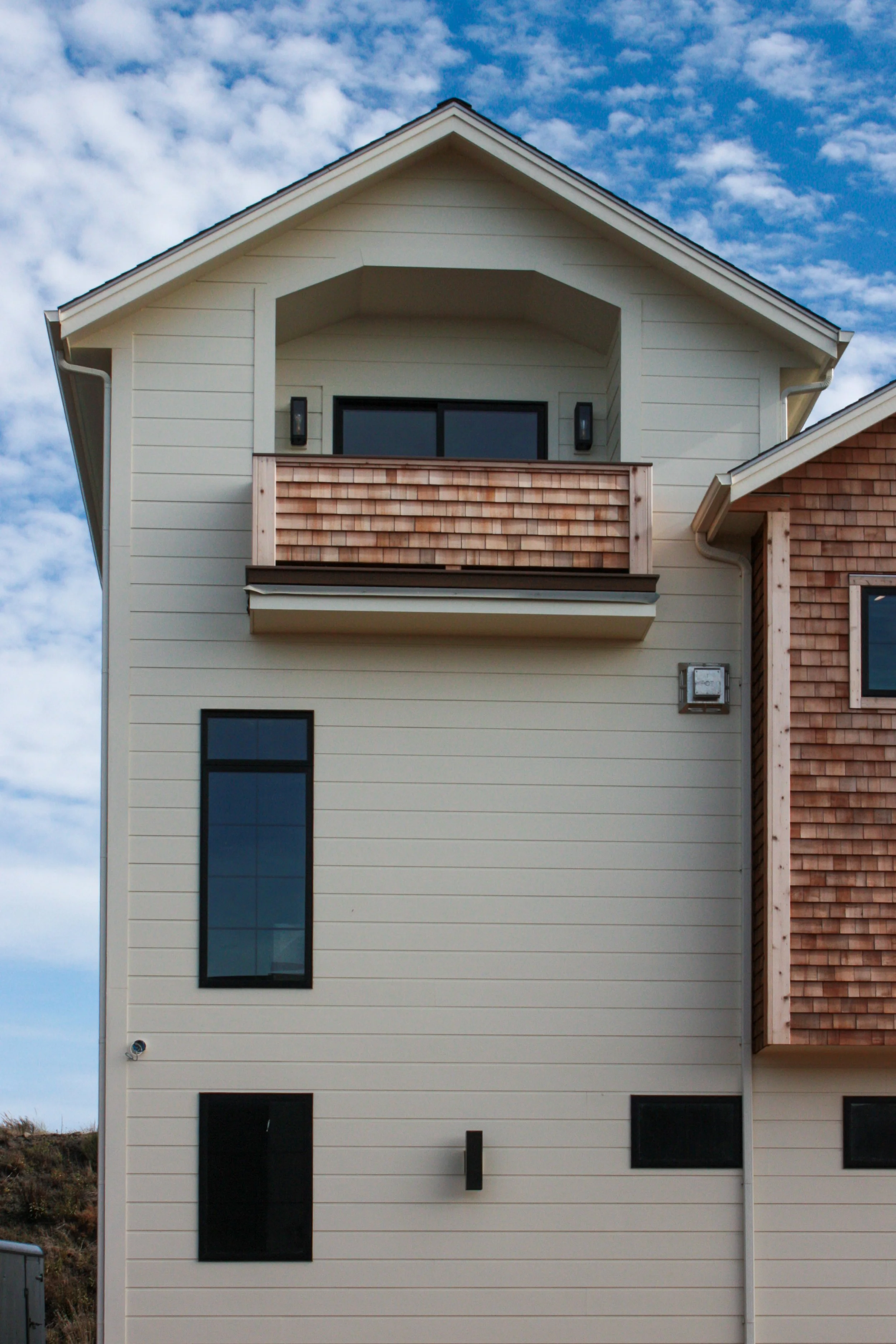 Front view of a modern multi-story house with beige siding, a balcony with wooden railing, and black-framed windows under a partly cloudy sky.