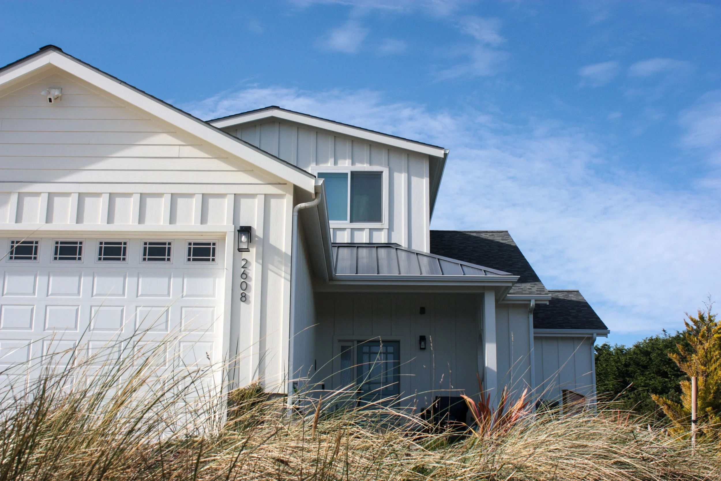 Modern white house with multiple gable roofs, black outdoor light fixture, gray window, and a partially covered entrance, surrounded by tall dry grass under a blue sky.