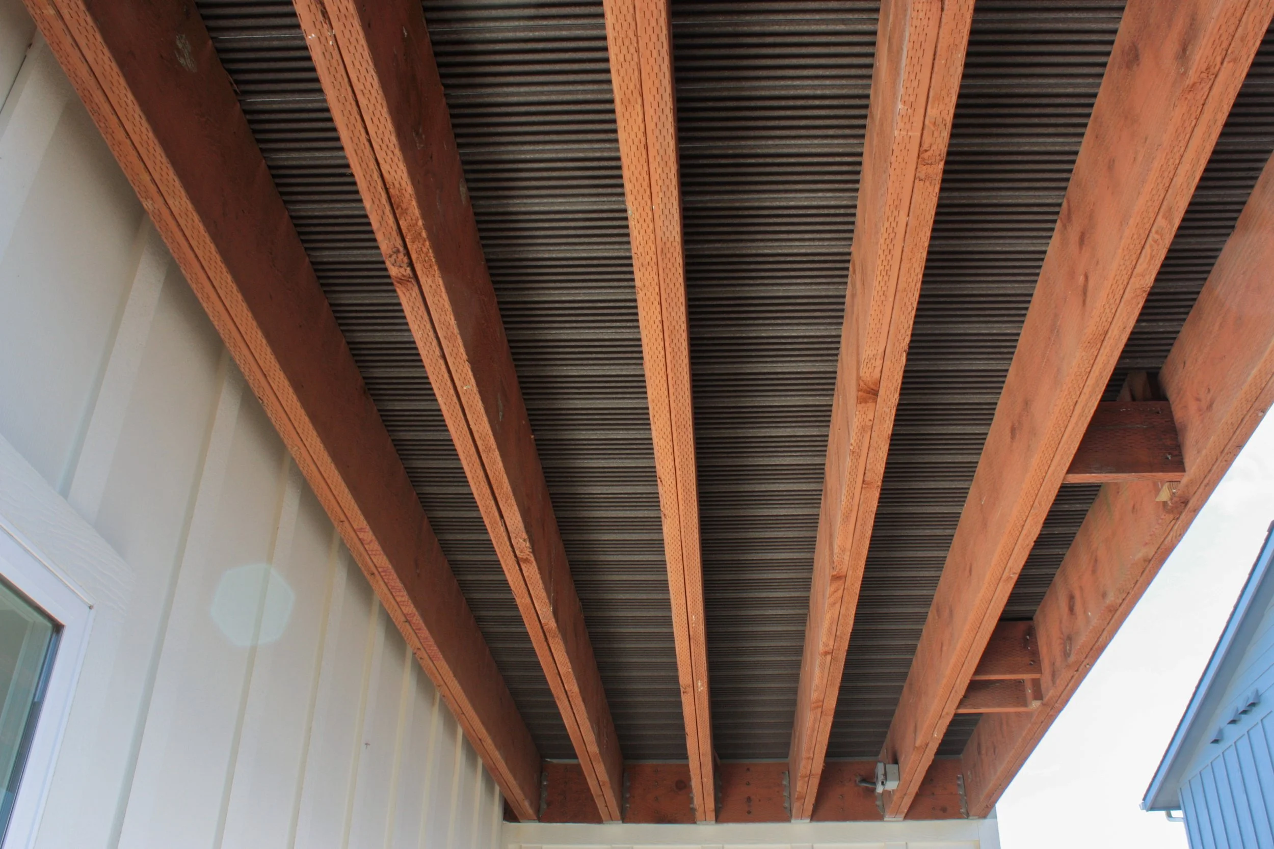 Underside of a roof with wooden beams and metal roofing sheets, attached to a light-colored wall with a window.