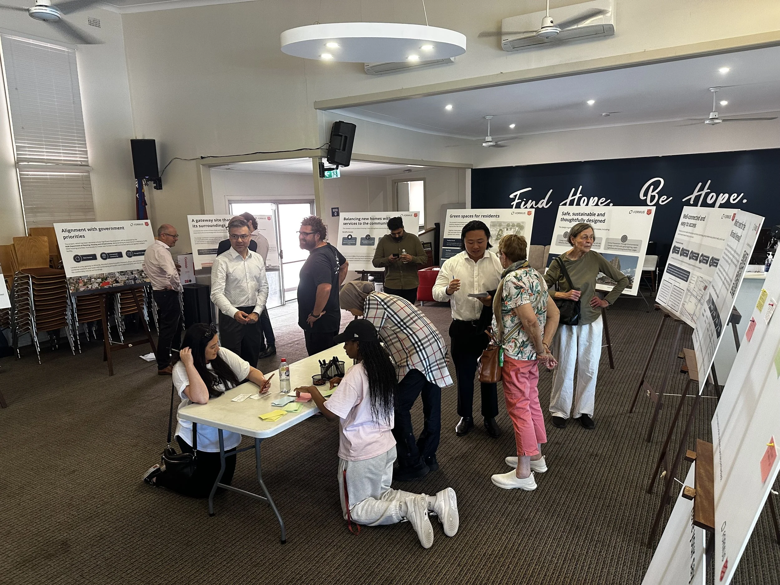 Display and discuss community session for residential development project. Participants writing feedback at a table surrounded by display boards, engaging in local planning and stakeholder consultation.