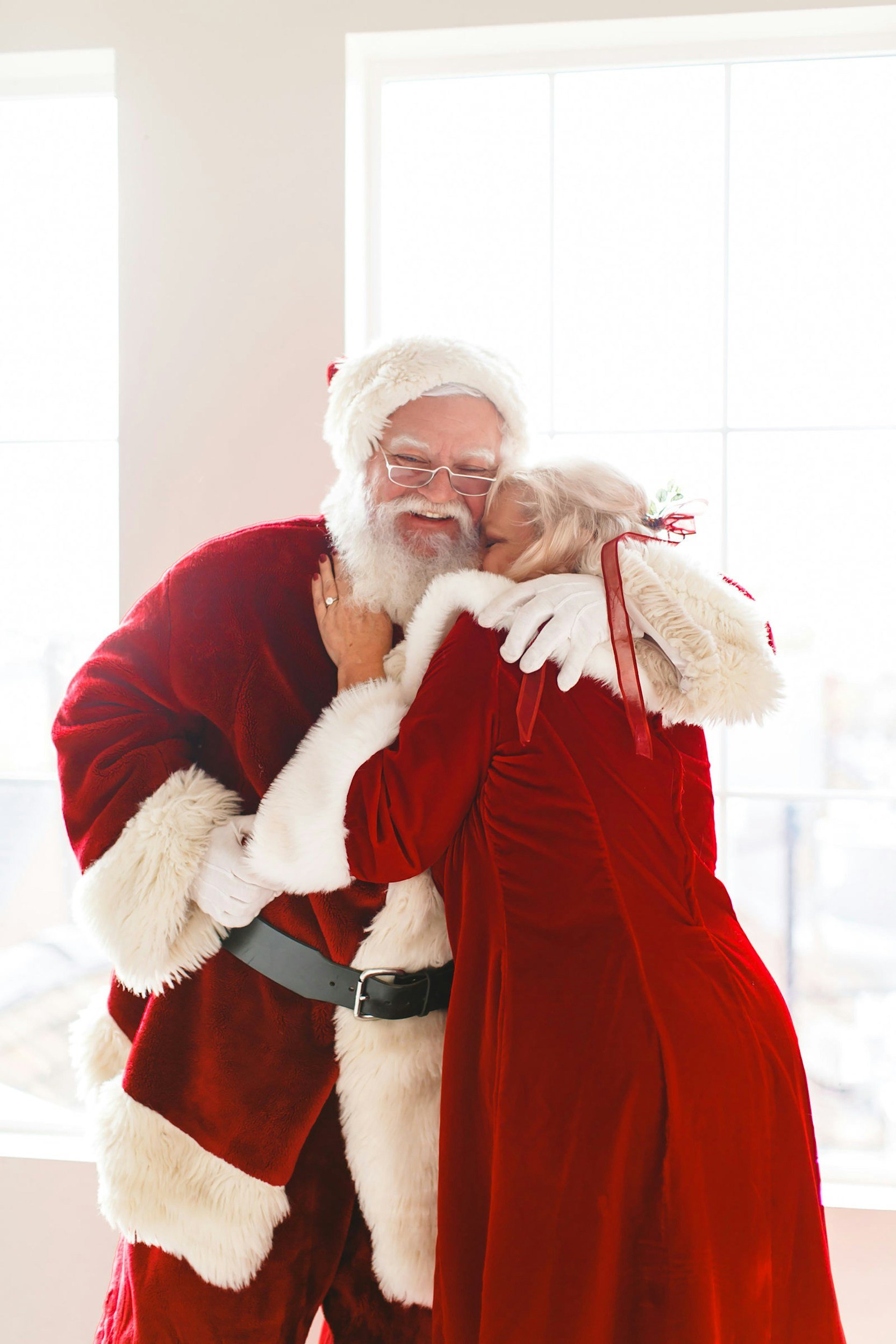 Santa Claus hugging a woman in a red dress, inside a bright room near a window.