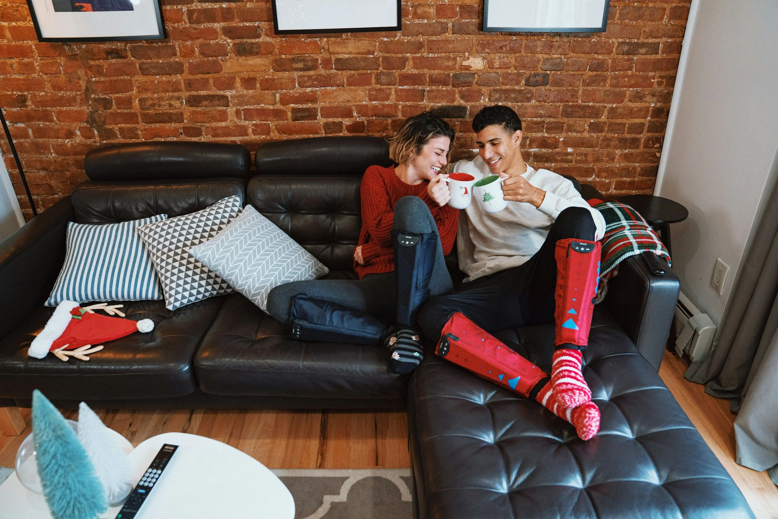 A young couple sitting on a leather sectional couch with a brick wall behind them, smiling and holding mugs, celebrating Christmas with holiday socks & decorations in the living room. Image is used for our CouplesTherapy.co Relationship Skills CLASS.