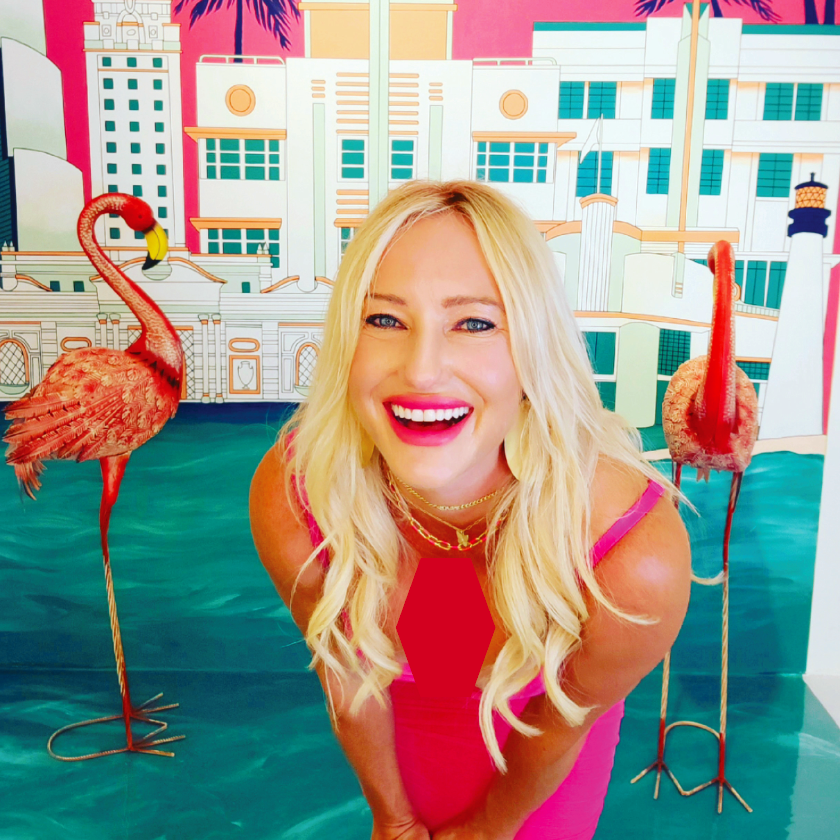 A smiling blonde woman in a pink dress, standing in front of a colorful Venice-themed backdrop with two flamingo statues and pink and teal water.