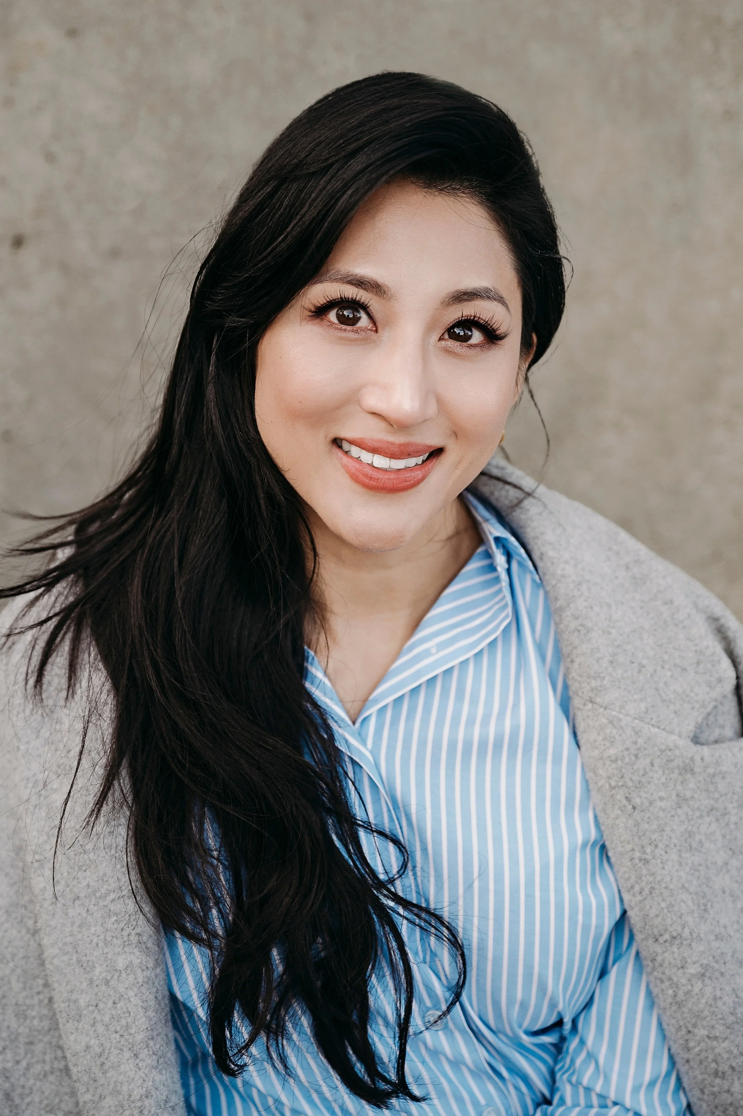 A woman with long dark hair, smiling, wearing a blue and white striped shirt and a gray coat, standing against a neutral background.