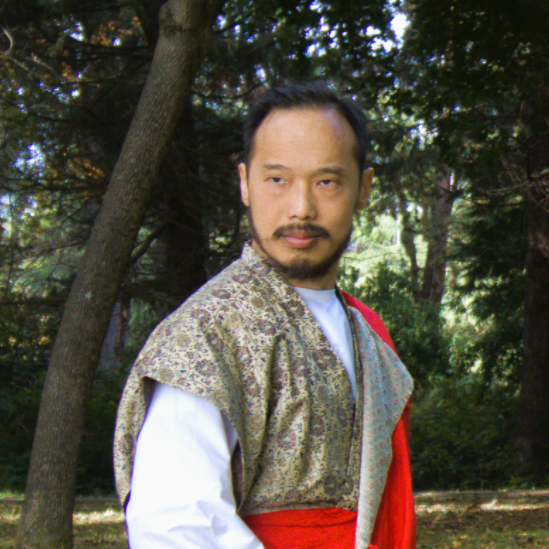 A man with short dark hair and facial hair standing outdoors among trees, wearing a patterned shawl over a white shirt and a red fabric draped over his shoulder.