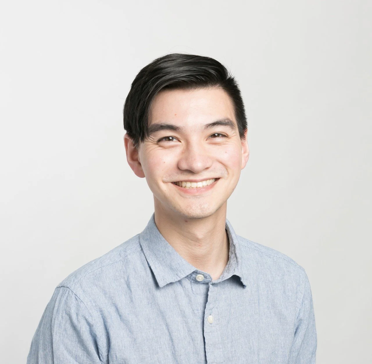Portrait of a young man with black hair wearing a light blue button-up shirt, smiling against a plain white background.