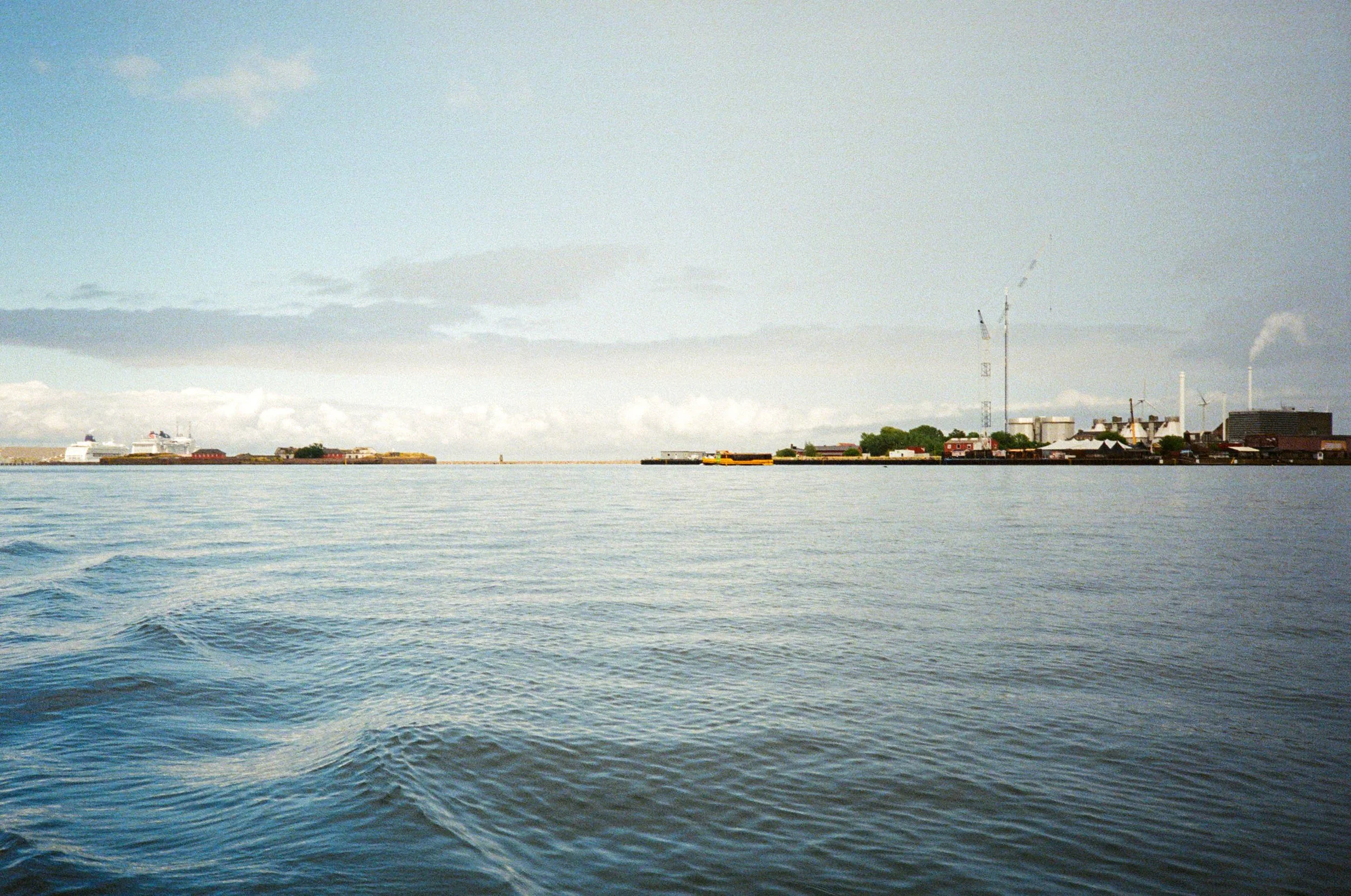 A view of a harbor with calm water, several ships docked, industrial buildings, and wind turbines in the distance under a partly cloudy sky.