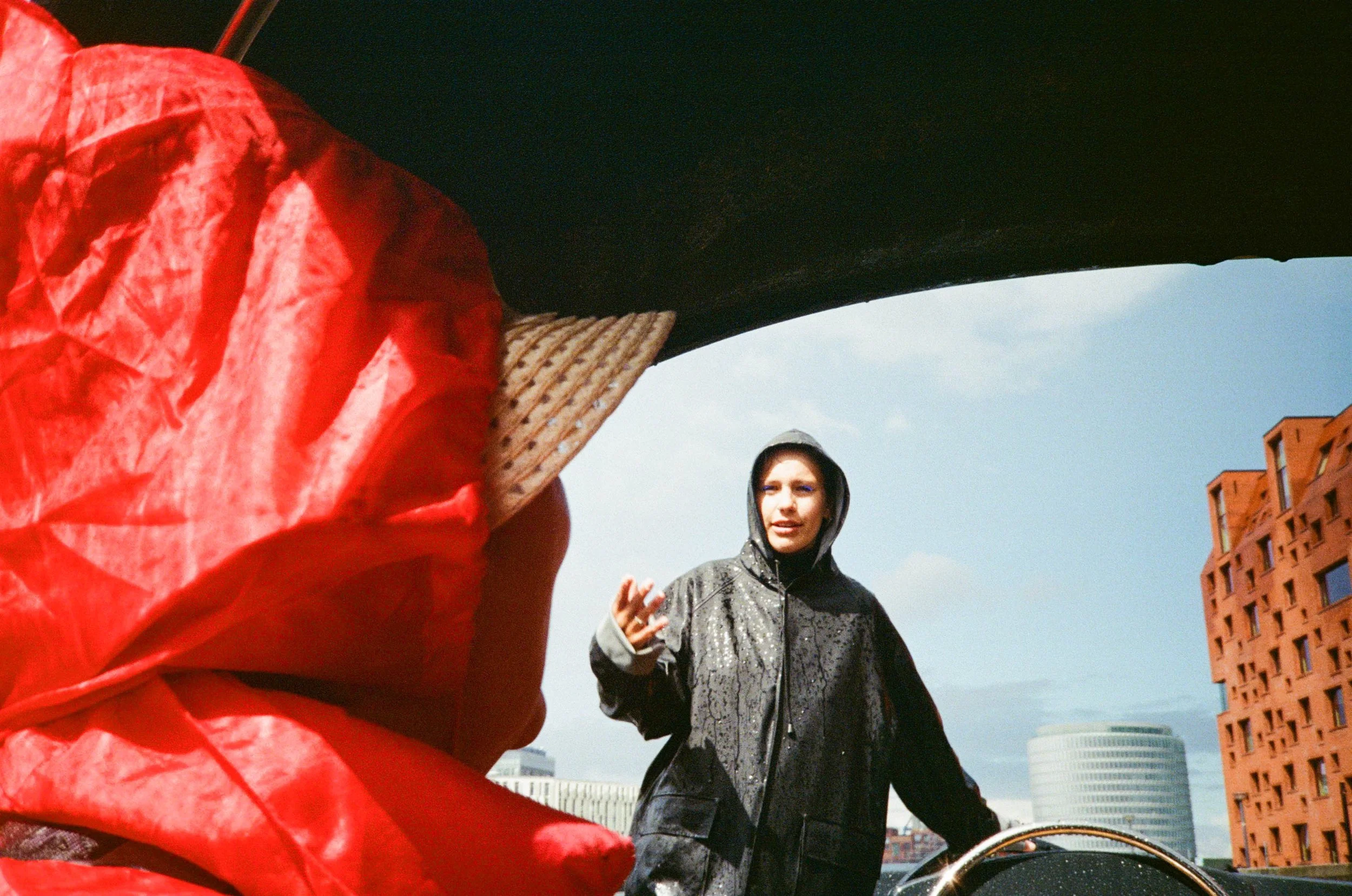 Image of a woman with dark hair, wearing a black shiny jacket with a hood, standing outdoors in an urban area, with a red brick building and a cloudy sky in the background. A person wearing a red jacket is in the foreground, partially blocking the vi