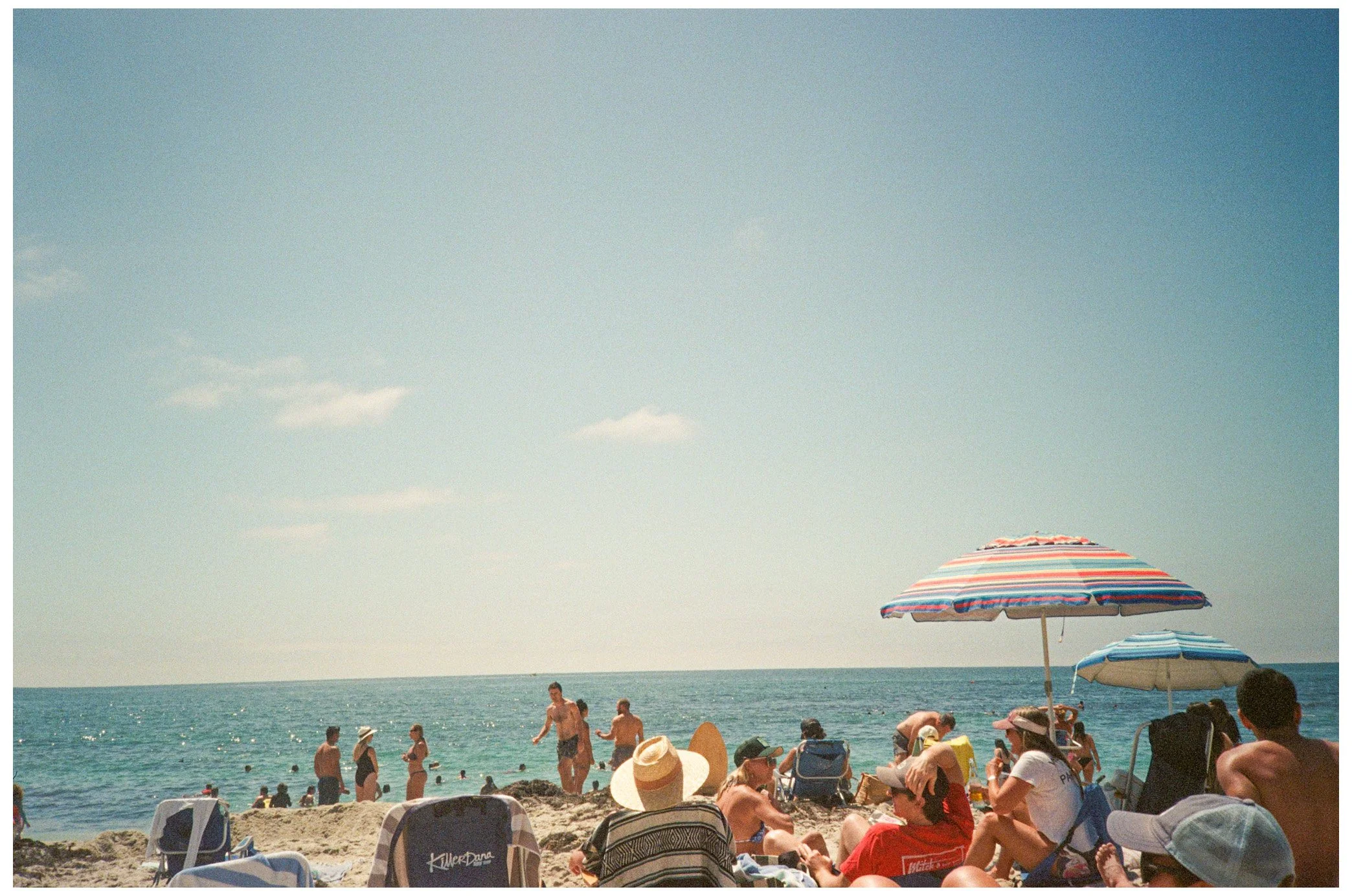 Crowded beach scene with people relaxing under umbrellas and swimming in the ocean on a sunny day.