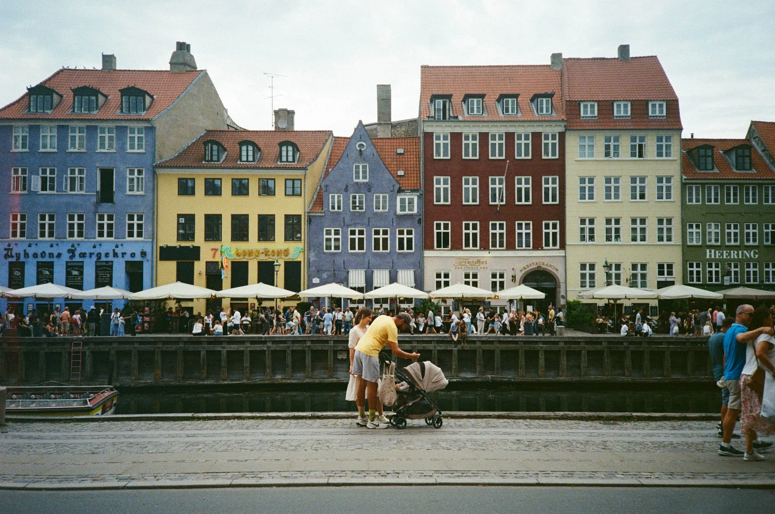 Colorful buildings along a waterfront with people walking and sitting outside under umbrellas.
