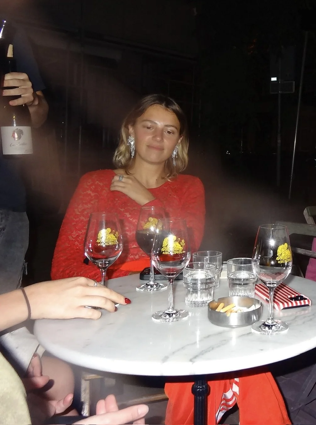 A woman in a red dress sitting at a marble table with several glasses of red wine, glasses of water, a small bowl with snacks, and a striped napkin, in a dimly lit setting, possibly a bar or restaurant.