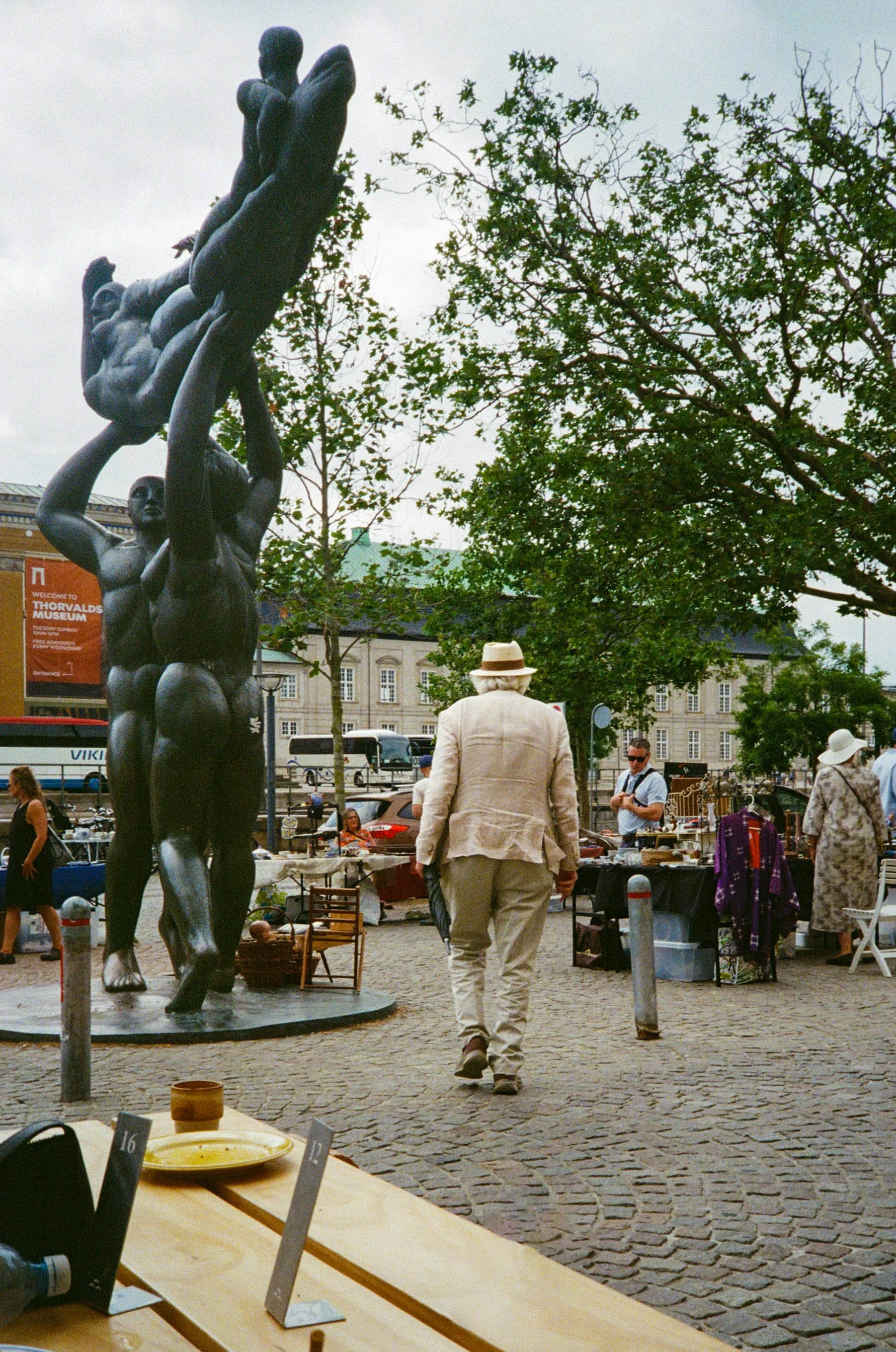 Street scene at an outdoor market with a bronze sculpture of a group of people holding hands. Several people browse the market stalls with various items. Historic buildings line the background, and a man with a hat walks away from the camera.