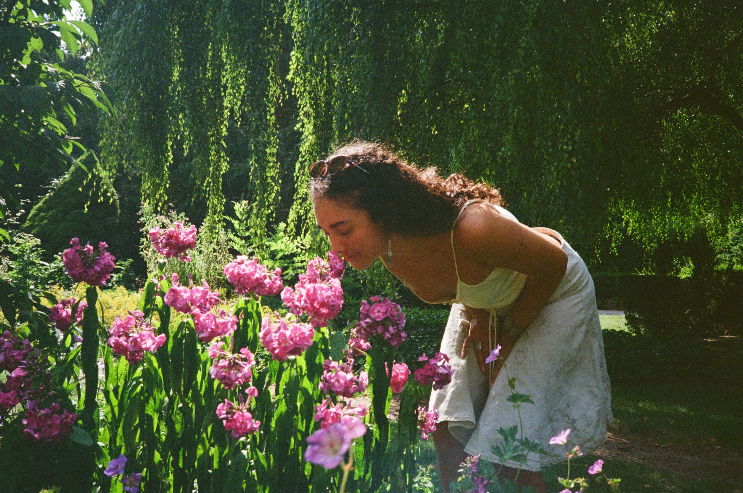 A woman with long dark hair, wearing sunglasses and a white sundress, is leaning forward and smelling pink flowers in a lush garden with green trees and hanging vines.