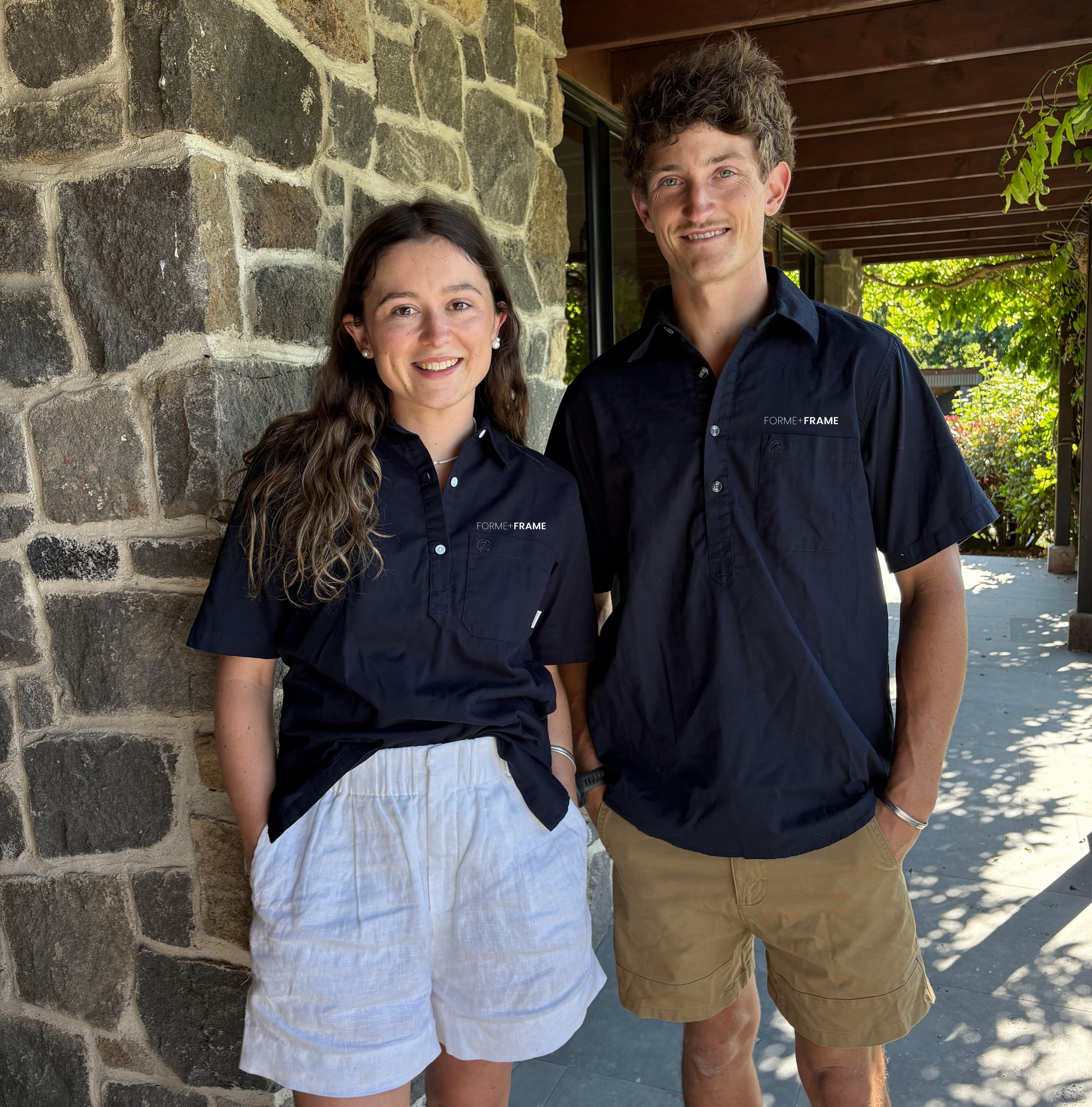 A young woman and a young man stand side by side outdoors near a stone wall, smiling at the camera. They are both wearing navy blue shirts with a logo that says 'FORME+FRAME.' The woman has long wavy brown hair and is wearing white shorts, while the man has curly brown hair and is wearing khaki shorts.