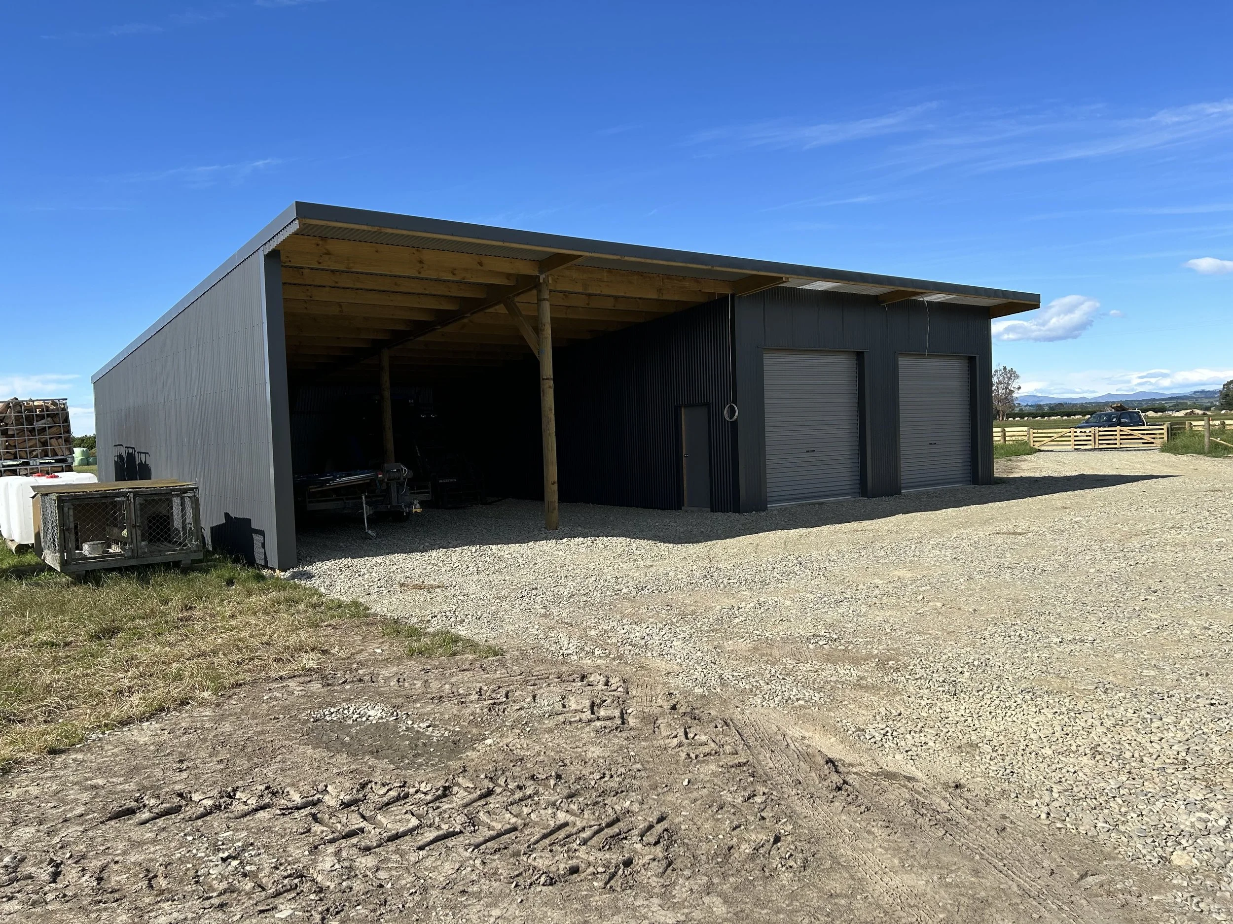 A large metal shed with two roll-up garage doors, a smaller side door, and a partially open section on the side with tools and equipment inside. Gravel driveway and open outdoor space with fencing and blue sky.