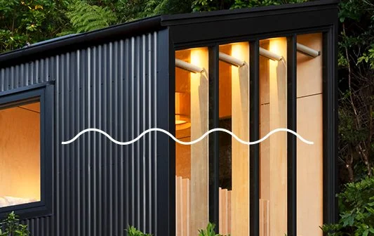 Modern tiny house with vertical wooden slats and black exterior siding, large window, surrounded by greenery.