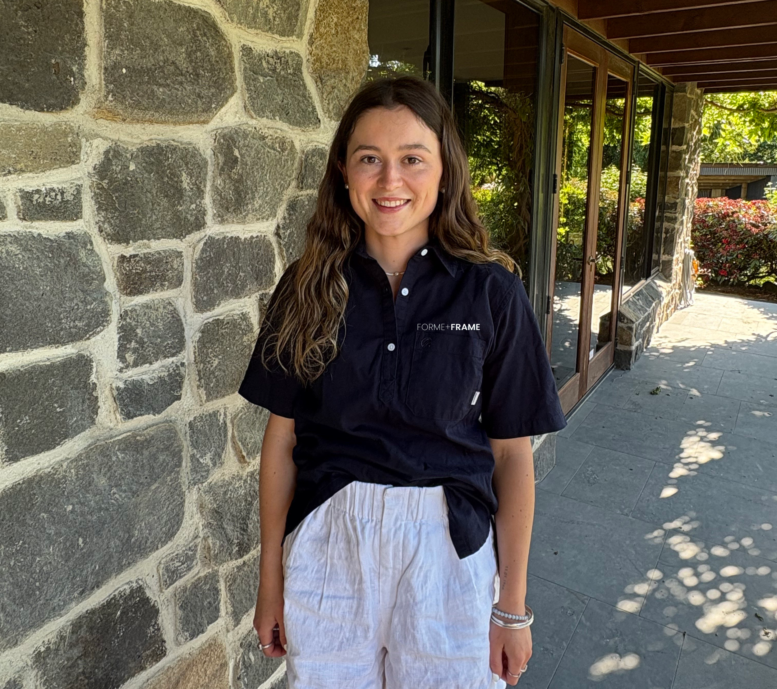 Young woman with long wavy brown hair smiling, wearing a black short-sleeved button-up shirt with white text, standing outside near a stone wall and glass windows, with greenery in the background.
