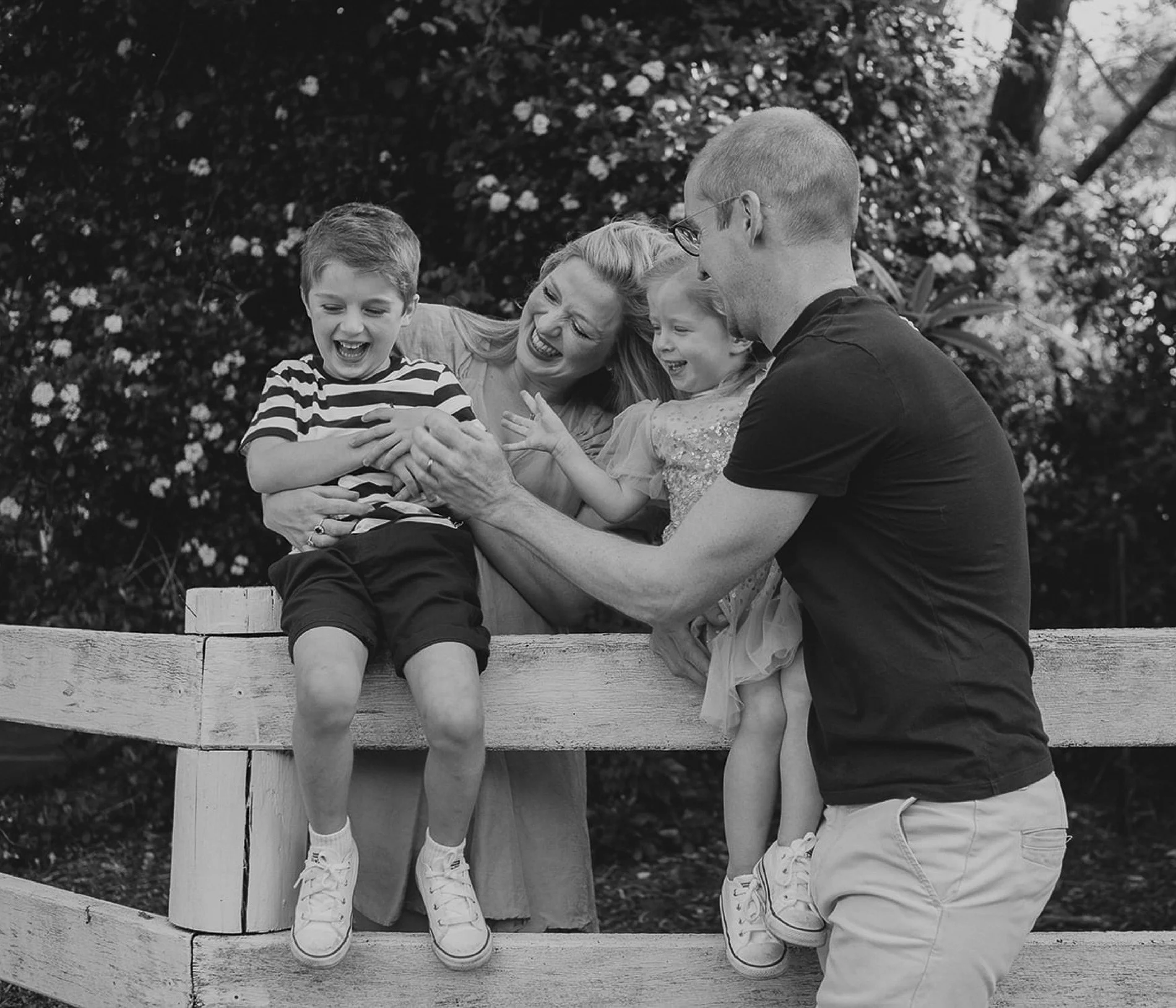 A happy family of four, including a mother, father, young boy, and young girl, enjoying a moment outside on a wooden fence, laughing and playing together.