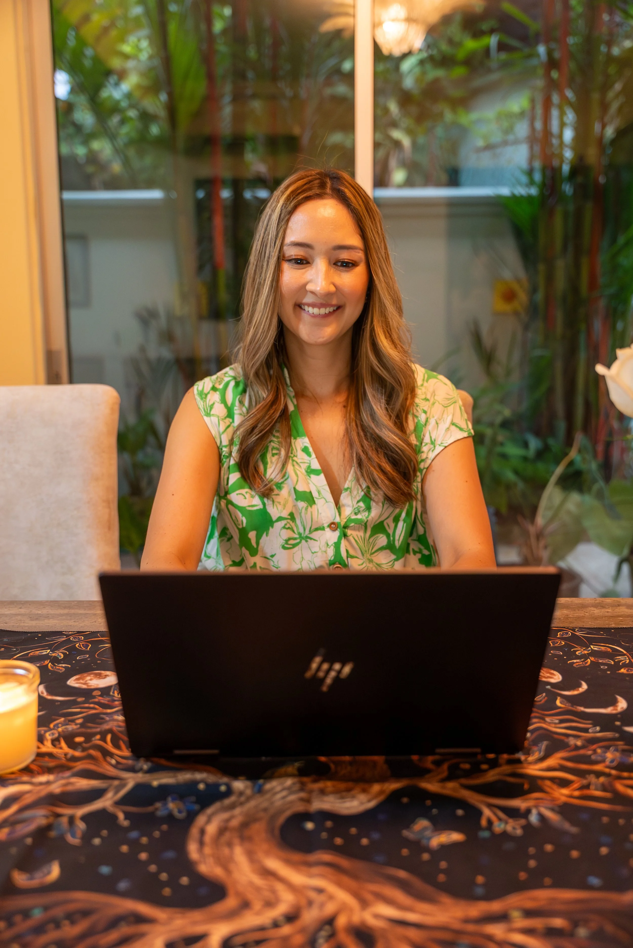A woman with long, wavy brown hair sitting at a table, looking at her laptop and smiling. The table has a black cloth with a celestial and tree design, and there is a lit candle on the left side. She is indoors with large glass windows behind her, showing green plants outside.