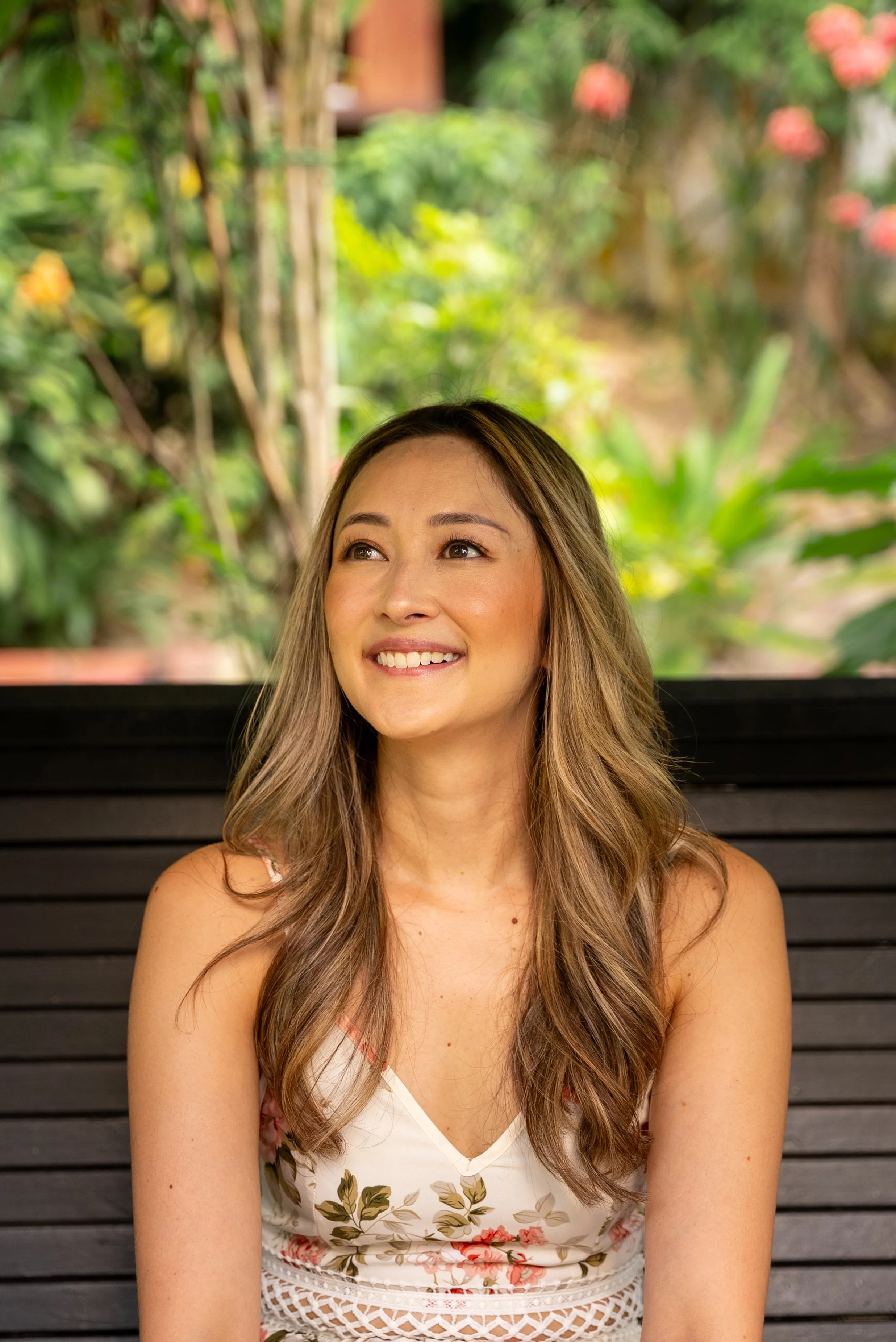 A woman with long, wavy hair, smiling and looking slightly upward, sitting on a black bench outdoors with lush green and pink plants in the background.