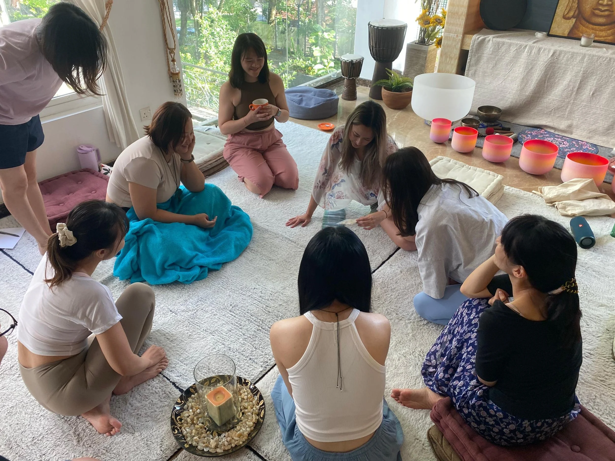 A group of women gather in a circle on a carpeted floor, engaged in a meditation or mindfulness activity in a bright room with large windows, plants, and decorative objects in the background.