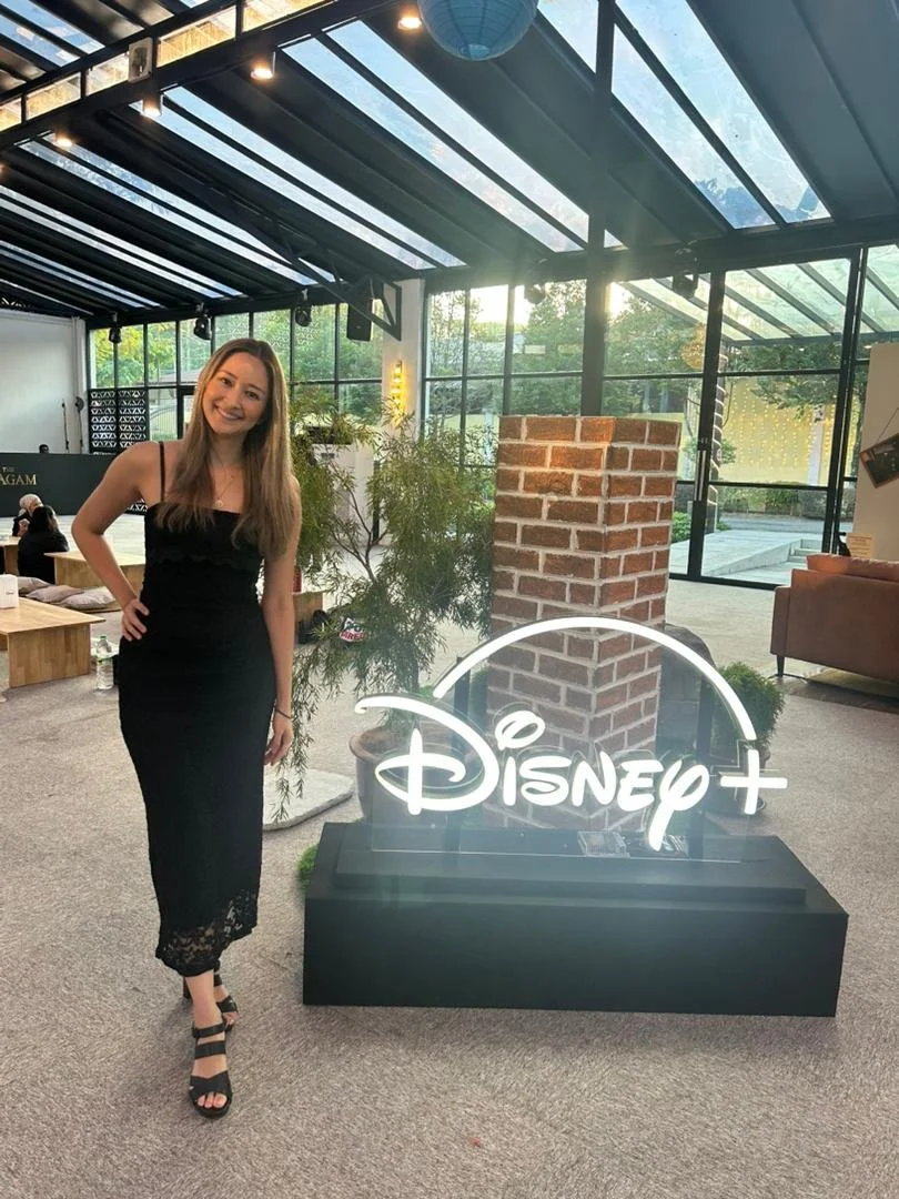A woman in a black dress and heels standing next to a Disney+ neon sign inside a modern glass-enclosed space with plants and brick decor.