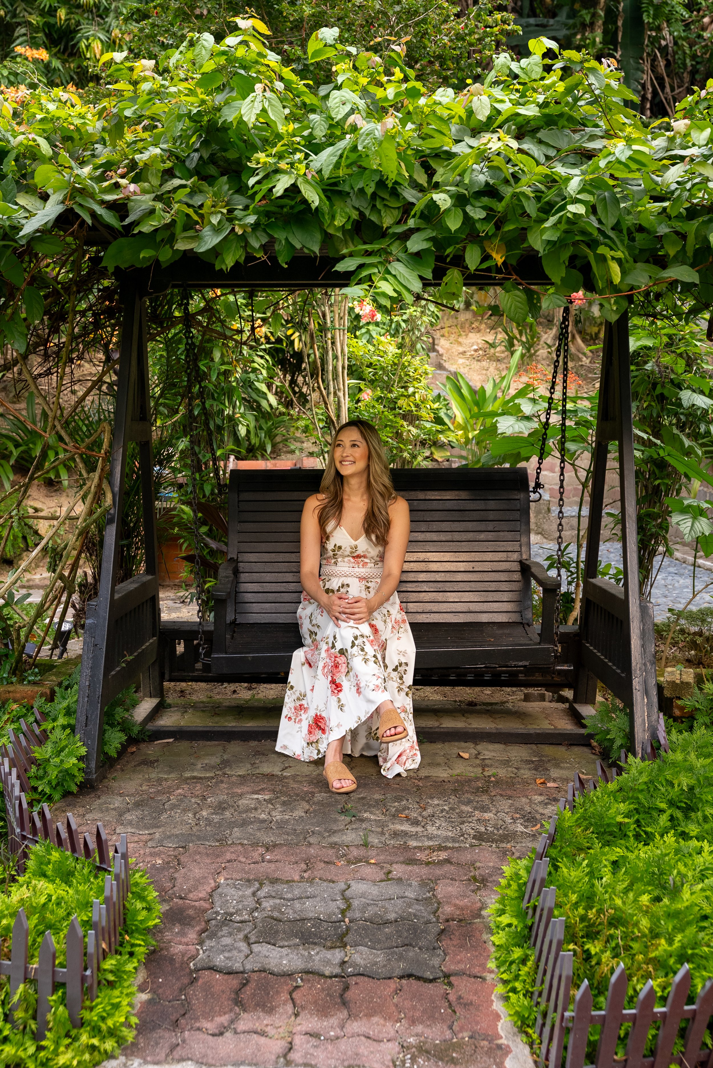 A woman in a floral dress sitting on a black outdoor swing with a canopy of green leaves above her in a lush garden.