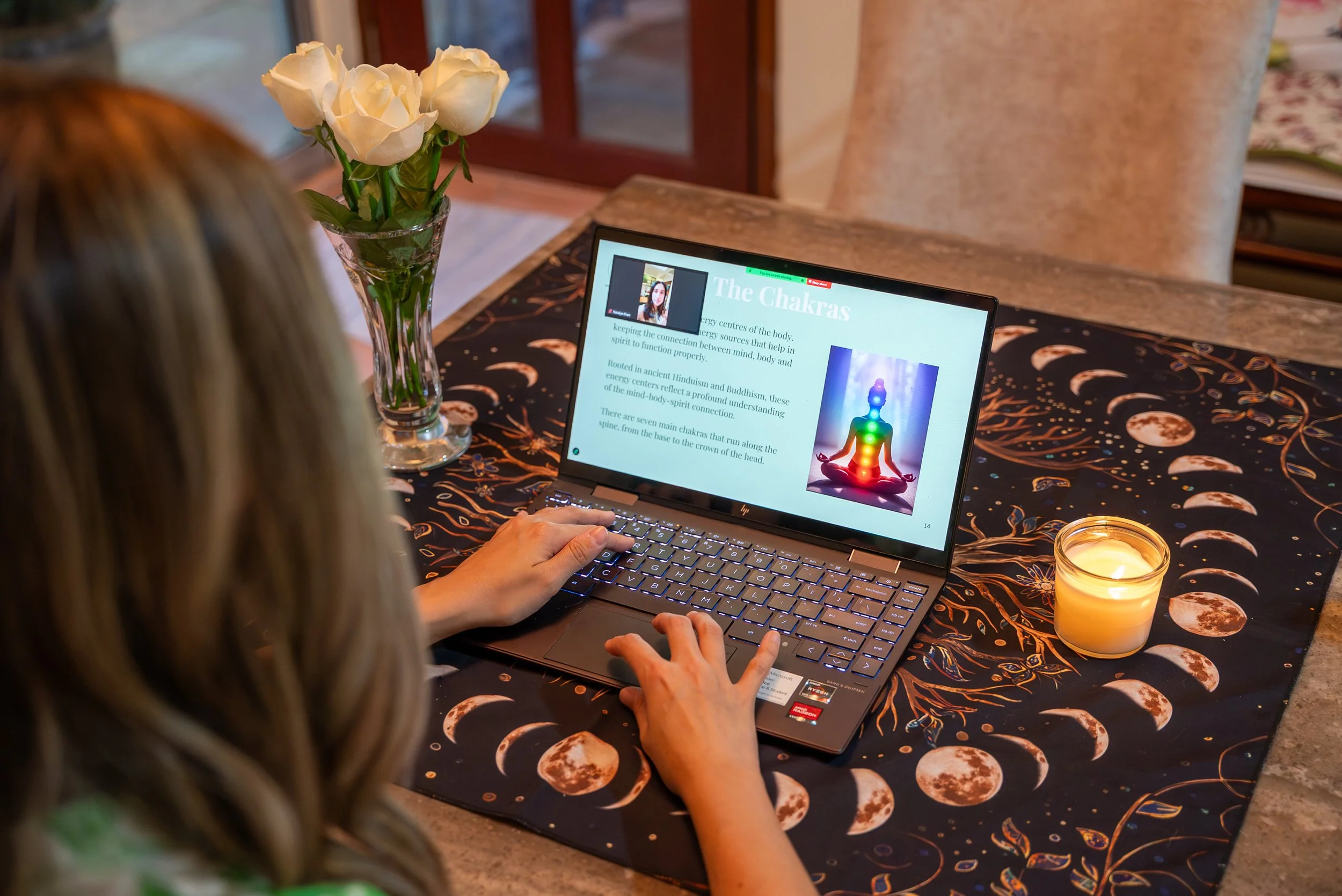 Person with long hair sitting at a dining table covered with a celestial-themed tablecloth, using a laptop to view an online presentation about the chakras. A vase of white roses, a lit candle, and a window are visible on the table.