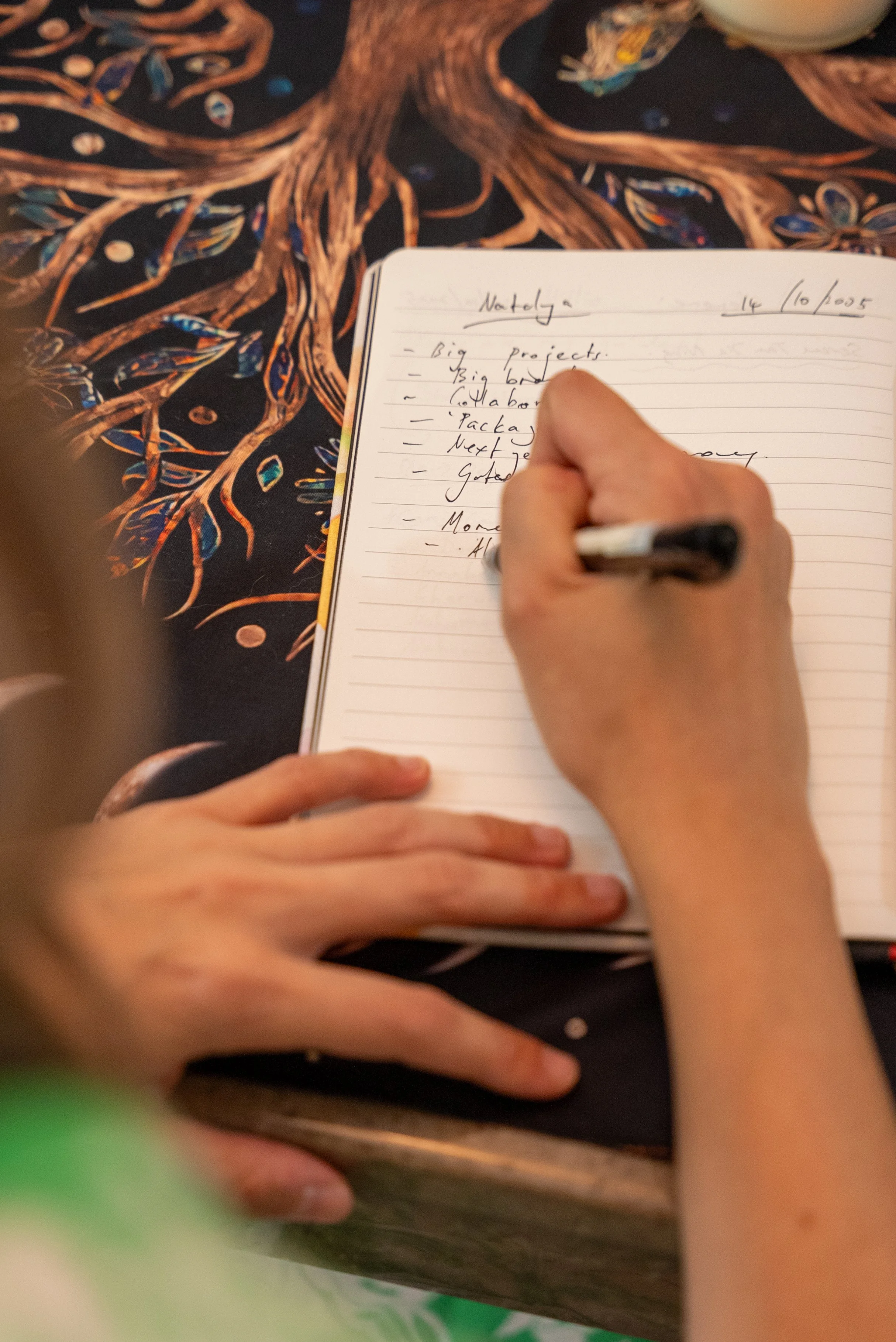 A person is writing notes in a notebook on a table with a dark, colorful, abstract tablecloth featuring a tree design.