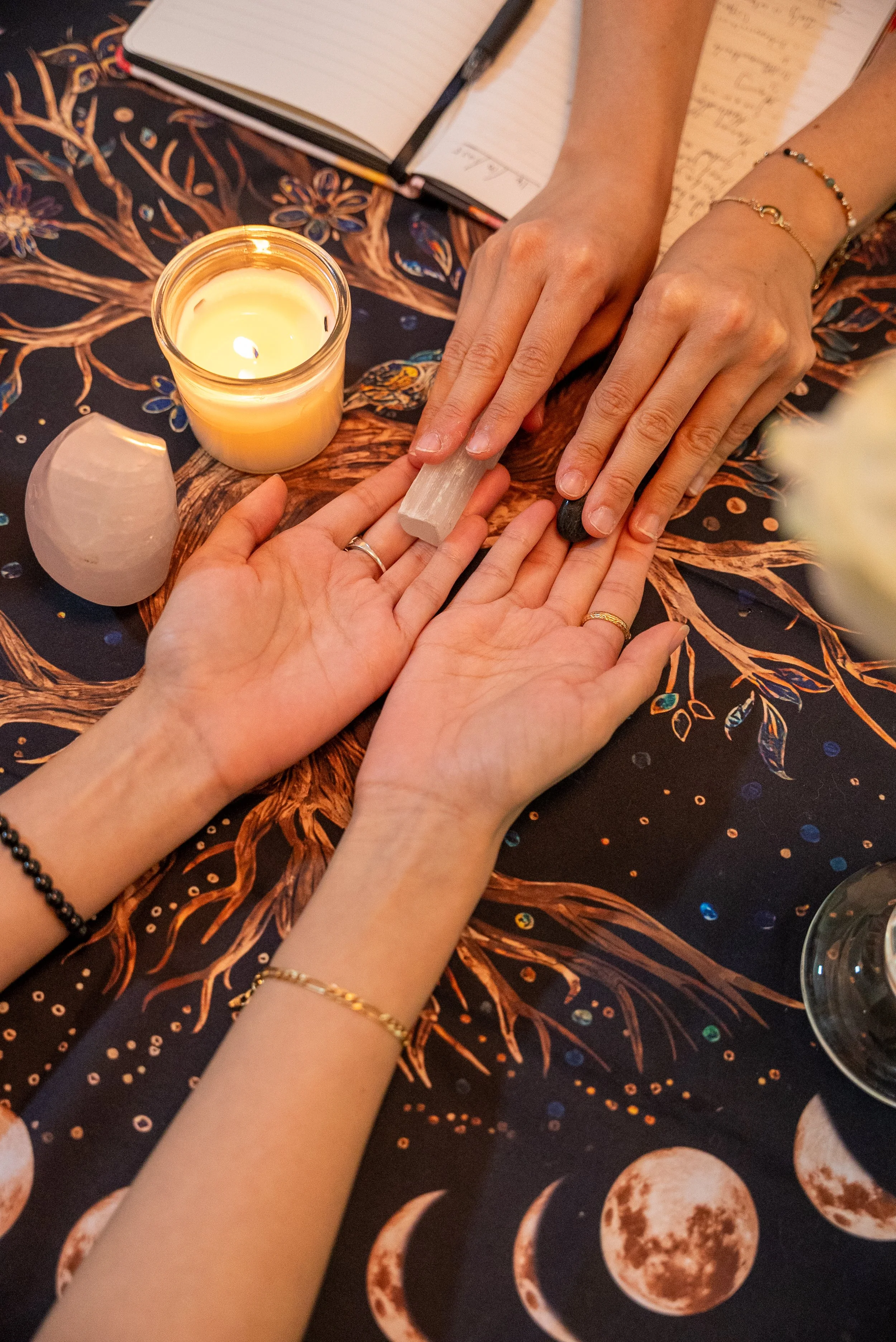 A person giving a manicure at a table with a dark celestial-themed tablecloth, a lit candle, and open notebooks.