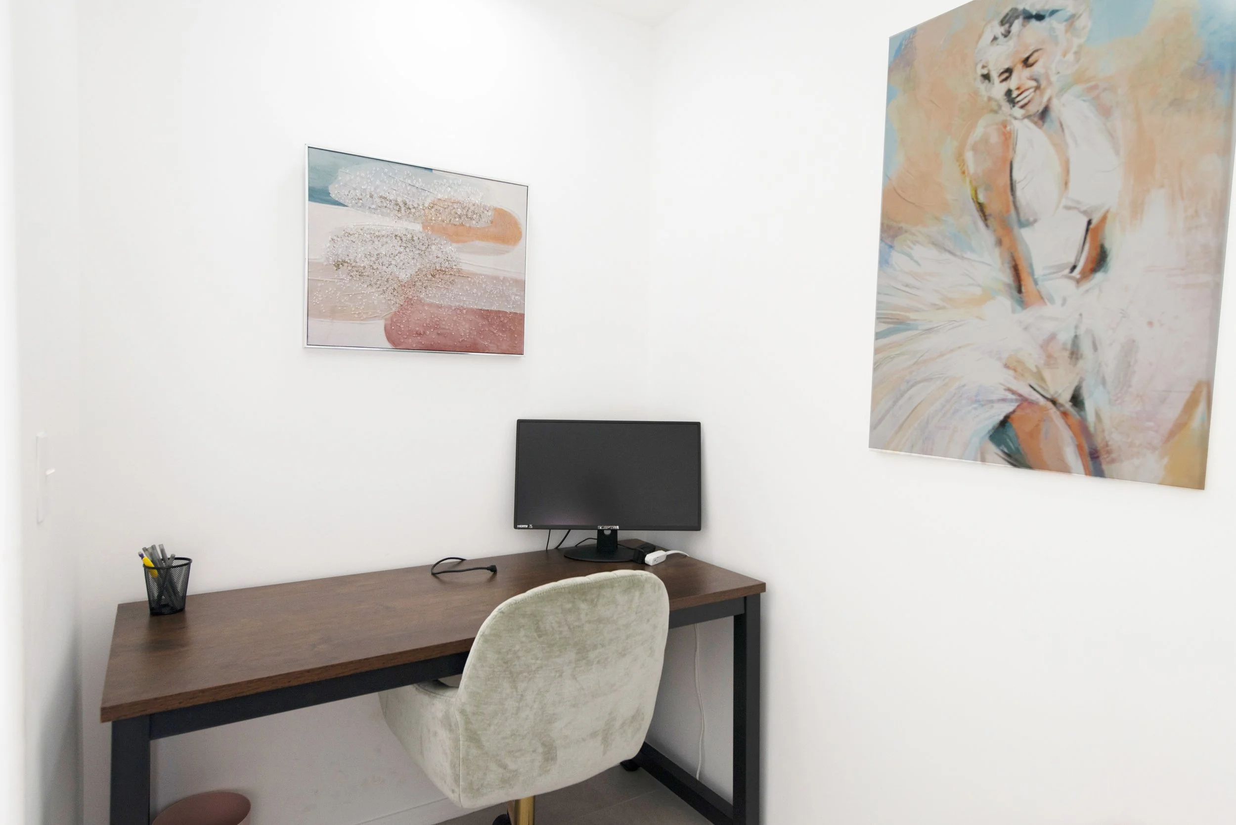 A small office space with a wooden desk, a beige upholstered chair, a computer monitor, a black pen holder with pens, and two paintings on the white walls—one abstract colored artwork and a portrait of a smiling woman in a dress.