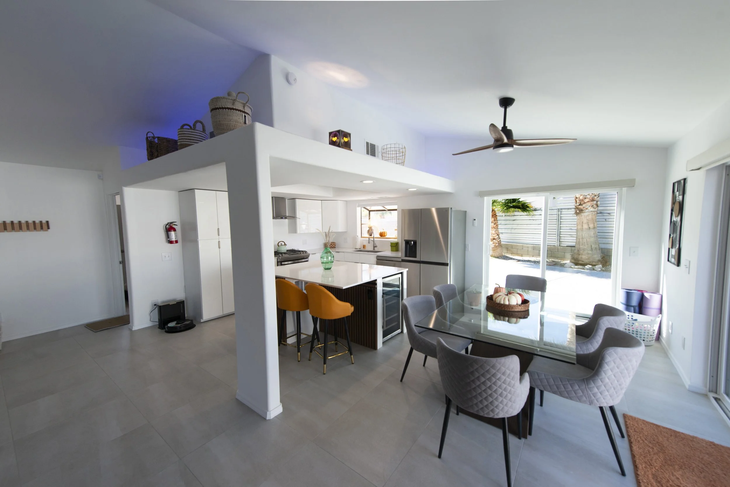 Modern open-concept kitchen and dining area with white cabinetry, stainless steel refrigerator, glass-top table, six gray upholstered chairs, and a view of the backyard with palm trees through sliding glass doors.