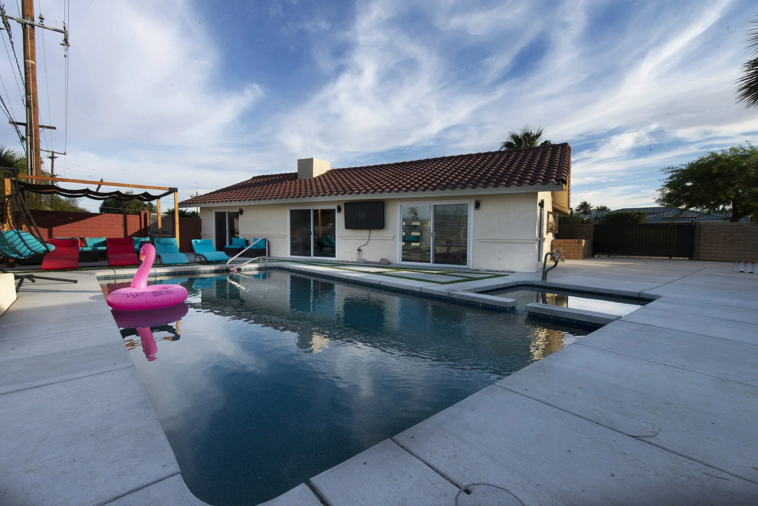 A backyard swimming pool with pink flamingo float, surrounded by poolside chairs and lounge area, house with sliding glass doors, and a partly cloudy sky.