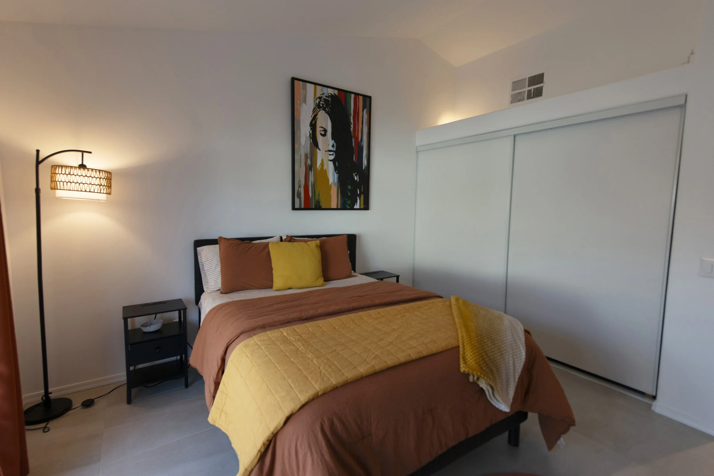 A bed with brown and yellow bedding, with three pillows in matching brown, yellow, and white. A black framed artwork of a woman hangs on the white wall above the bed. To the left, a black bedside table with a lamp and decorative bowl is visible. A sl