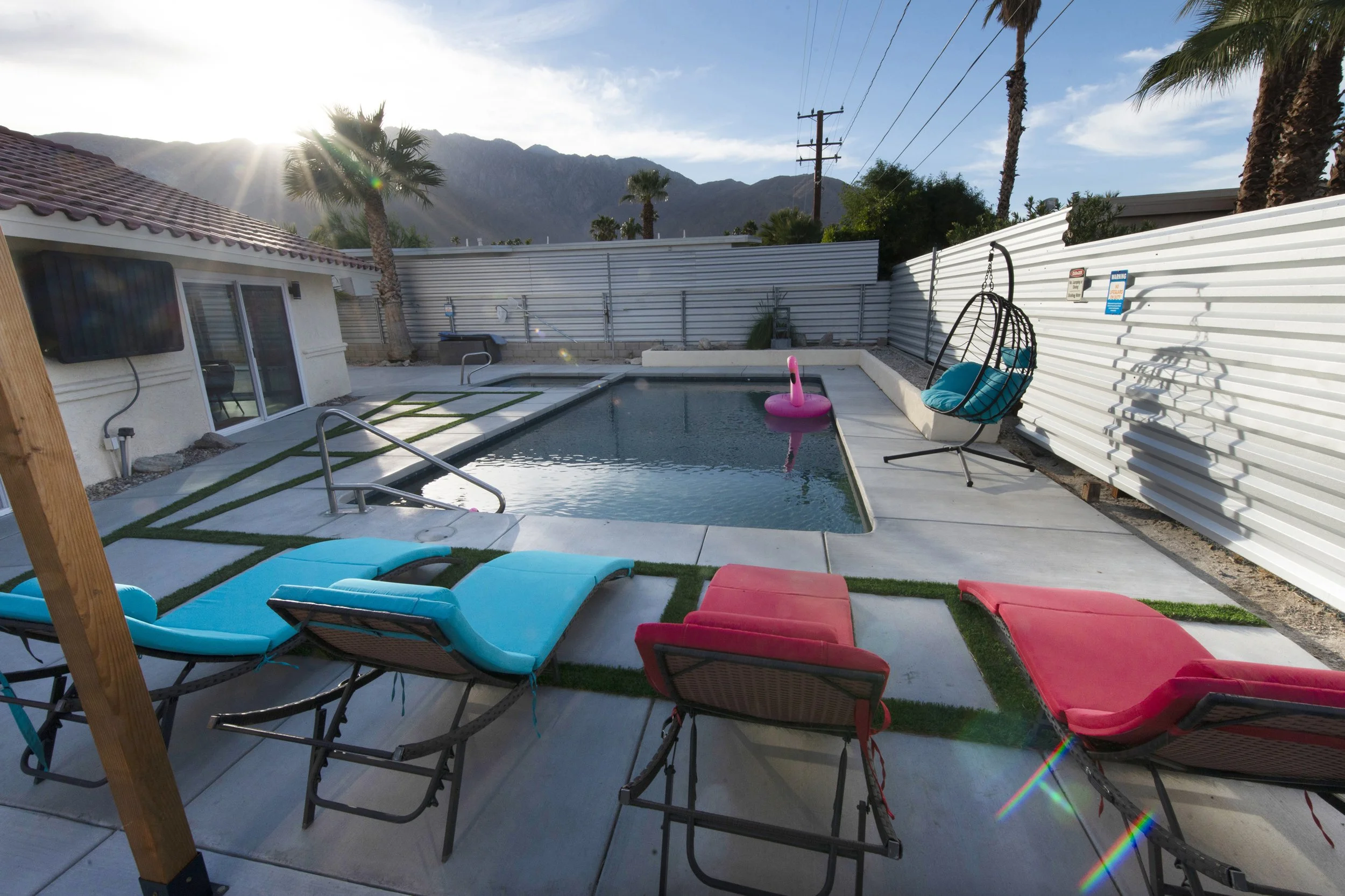 Backyard swimming pool area with lounge chairs, a hanging chair, a pink flamingo pool float, a small waterfall feature, and a corrugated metal fence, with mountain scenery in the background.