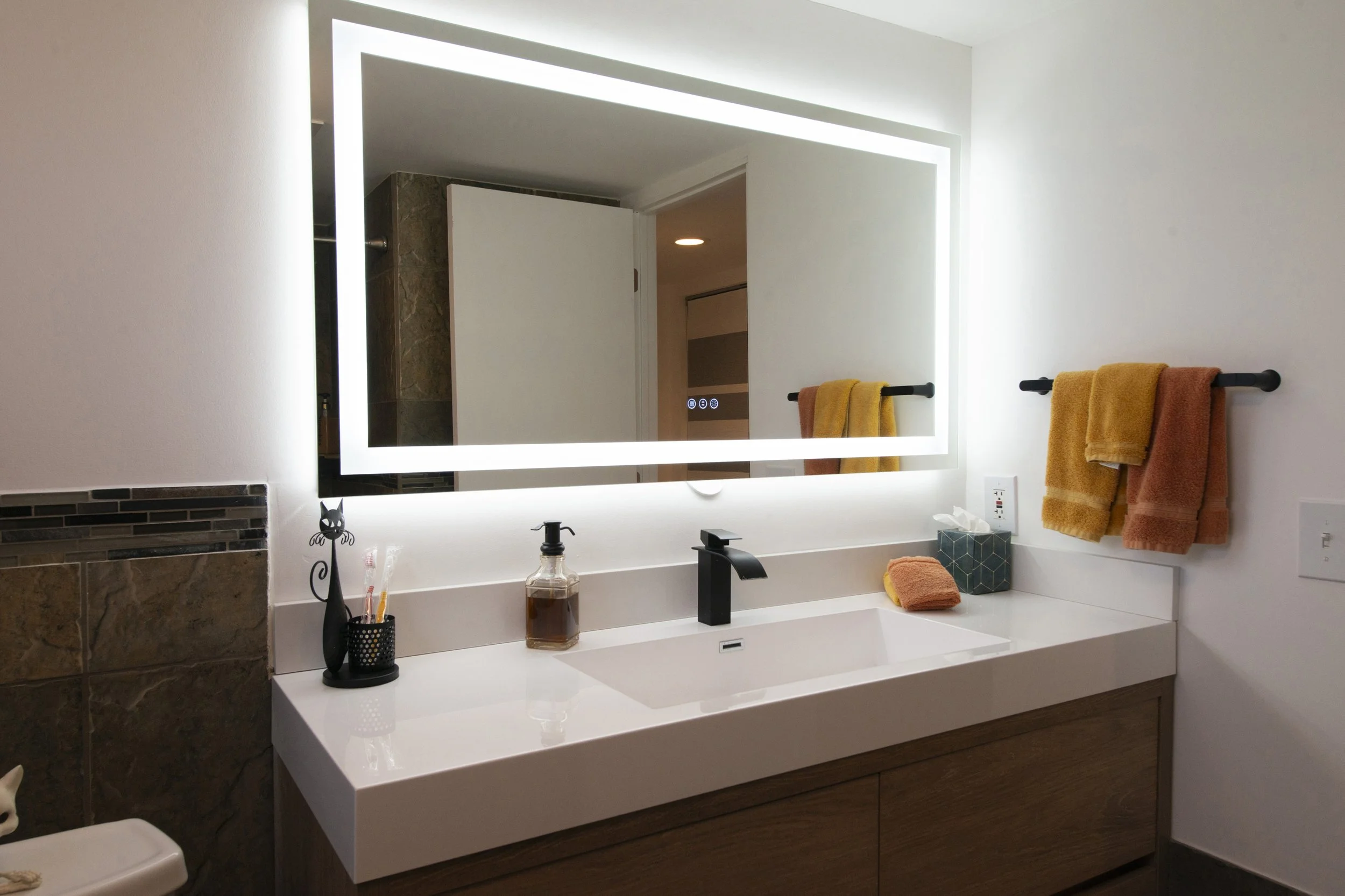 Modern bathroom vanity with a large backlit mirror, a black faucet, and beige and orange towels hanging on black towel bars