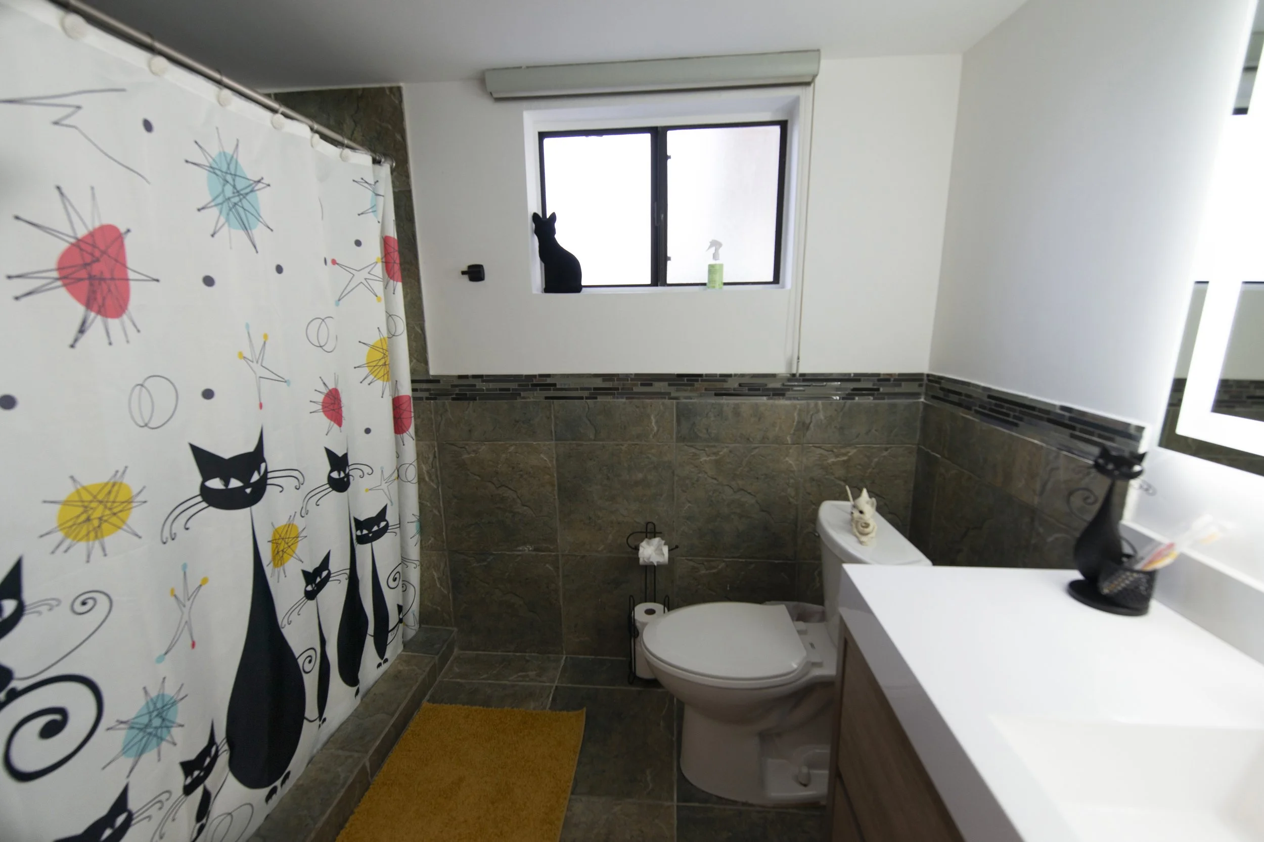 Bathroom with a shower curtain decorated with black cats and colorful abstract shapes, a window with a black cat silhouette, toilet, vanity with sink, and a yellow bath mat.