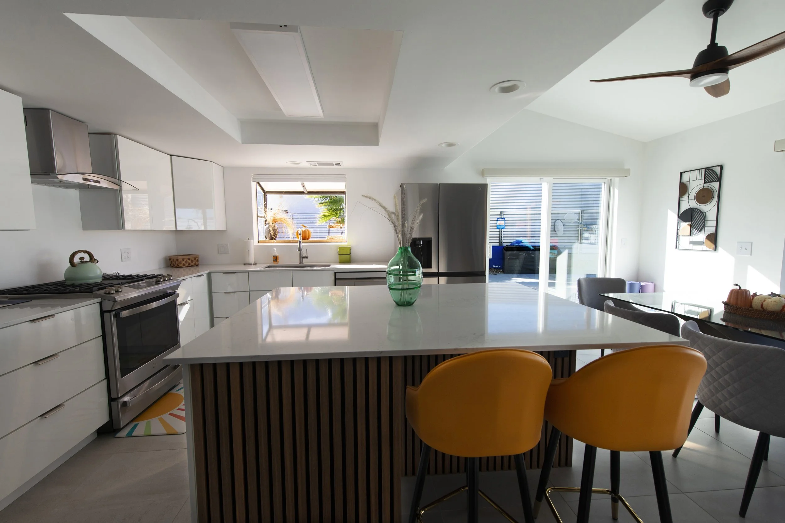 Modern kitchen with white cabinets, stainless steel appliances, a large island with a white countertop and yellow chairs, a sink by the window, and sliding glass doors leading outside.