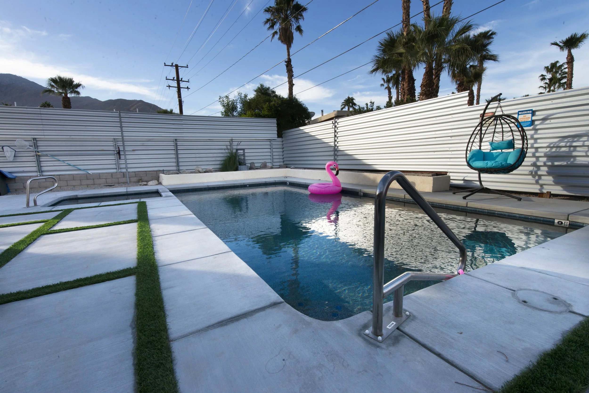 A backyard swimming pool with a pink inflatable swan, surrounded by a white concrete deck with green grass accents. There is a hanging blue seat on a stand and a white metal fence with palm trees in the background, under a partly cloudy sky.