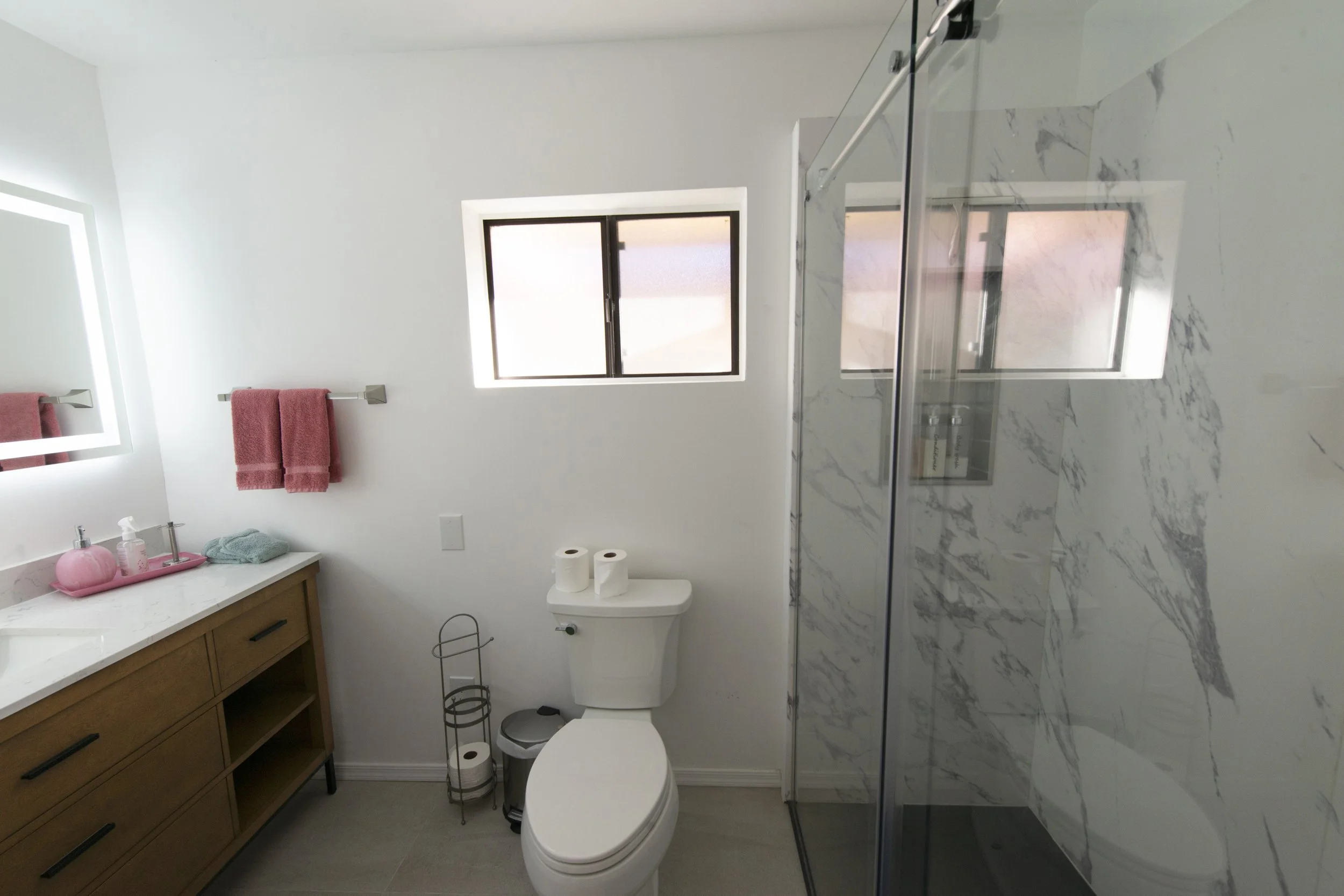 Modern bathroom with white walls, wooden vanity, pink towels, two rolls of toilet paper, a toilet, and a glass-enclosed shower with marble walls and a small window.