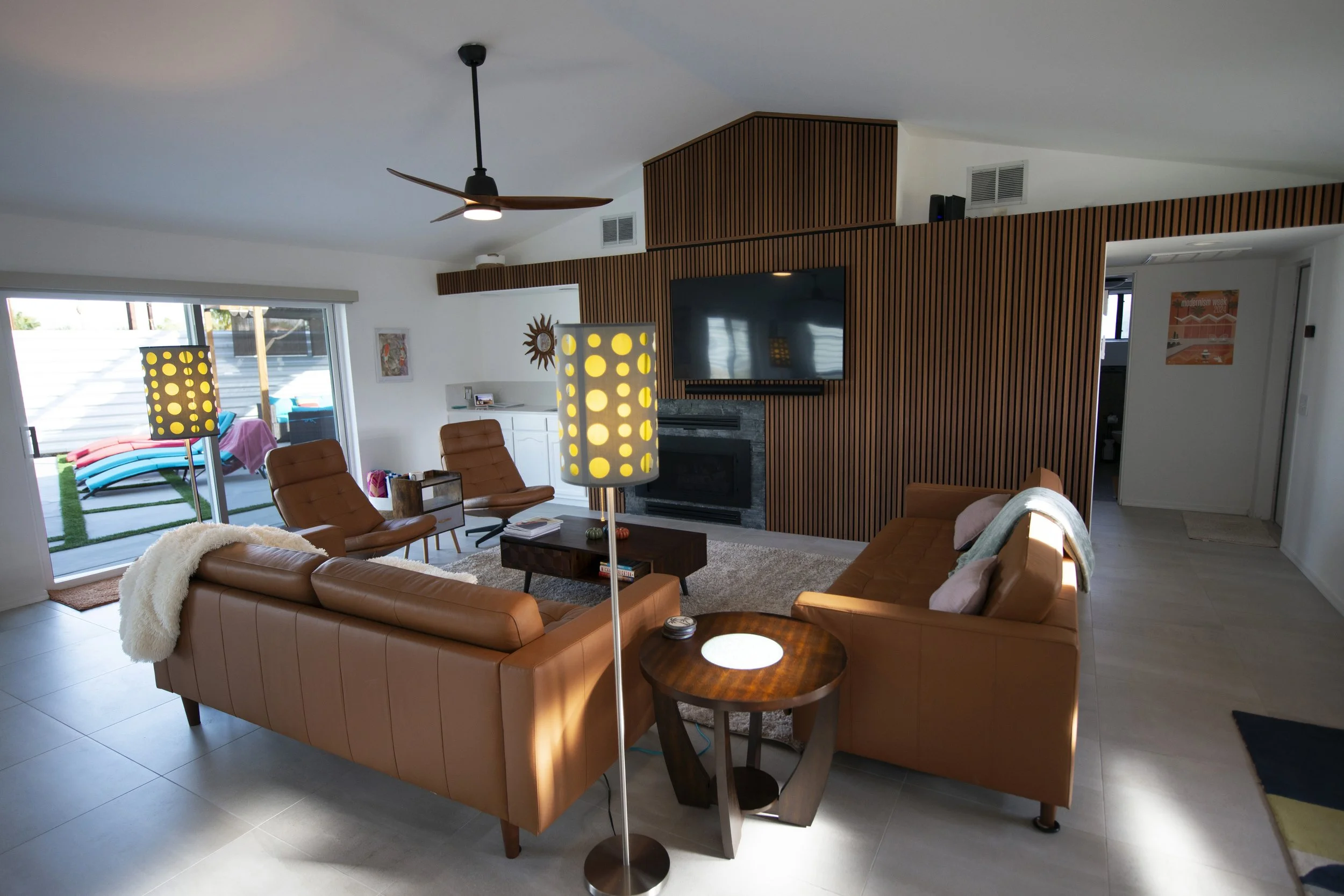 Living room with tan leather sofas, a wooden coffee table, a floor lamp with a yellow lampshade, a fireplace, and a wall-mounted TV, with a sliding door leading to pool and spa.